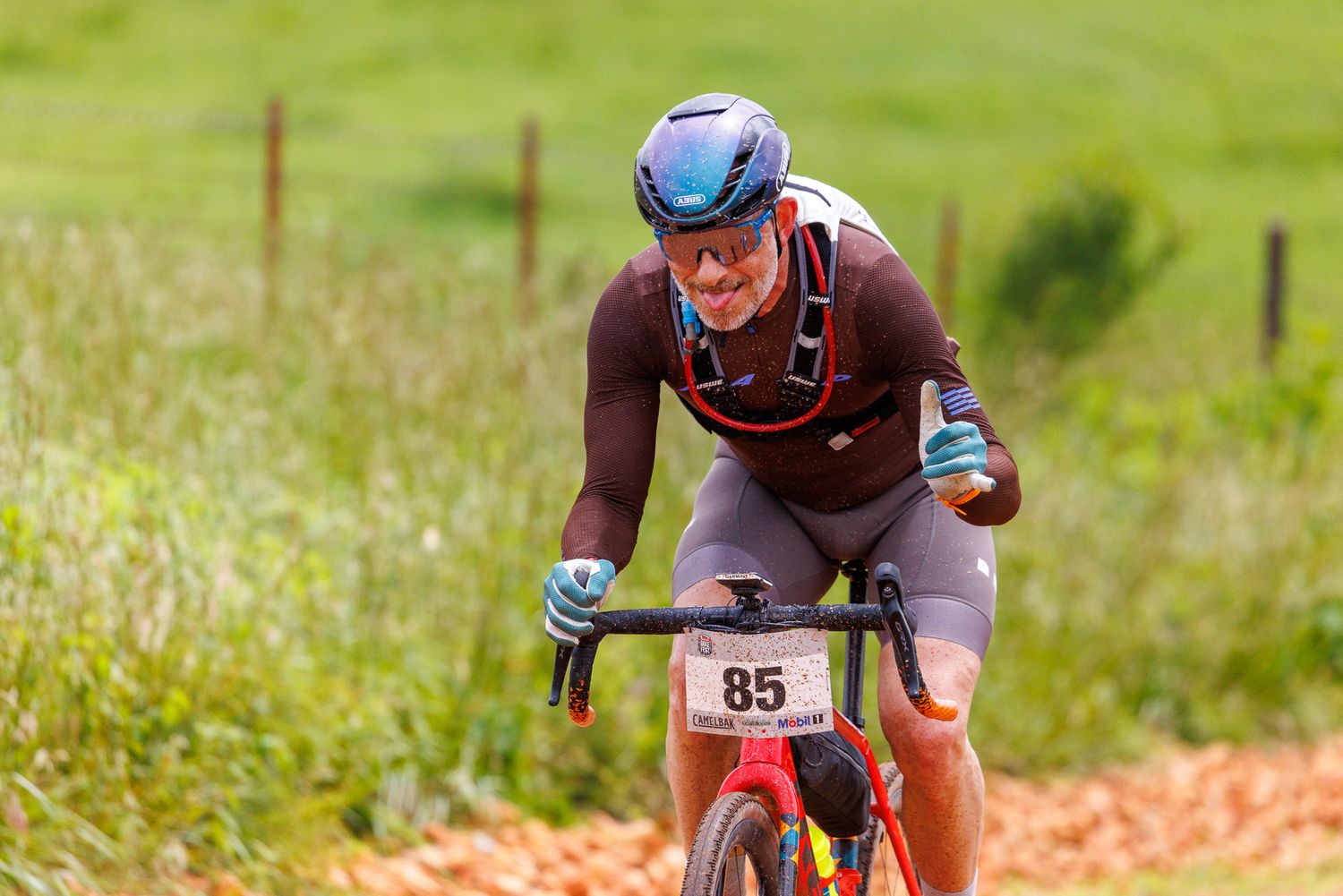 Determined gravel cyclist wearing race number 85 rides hard on a dirt road through green fields.