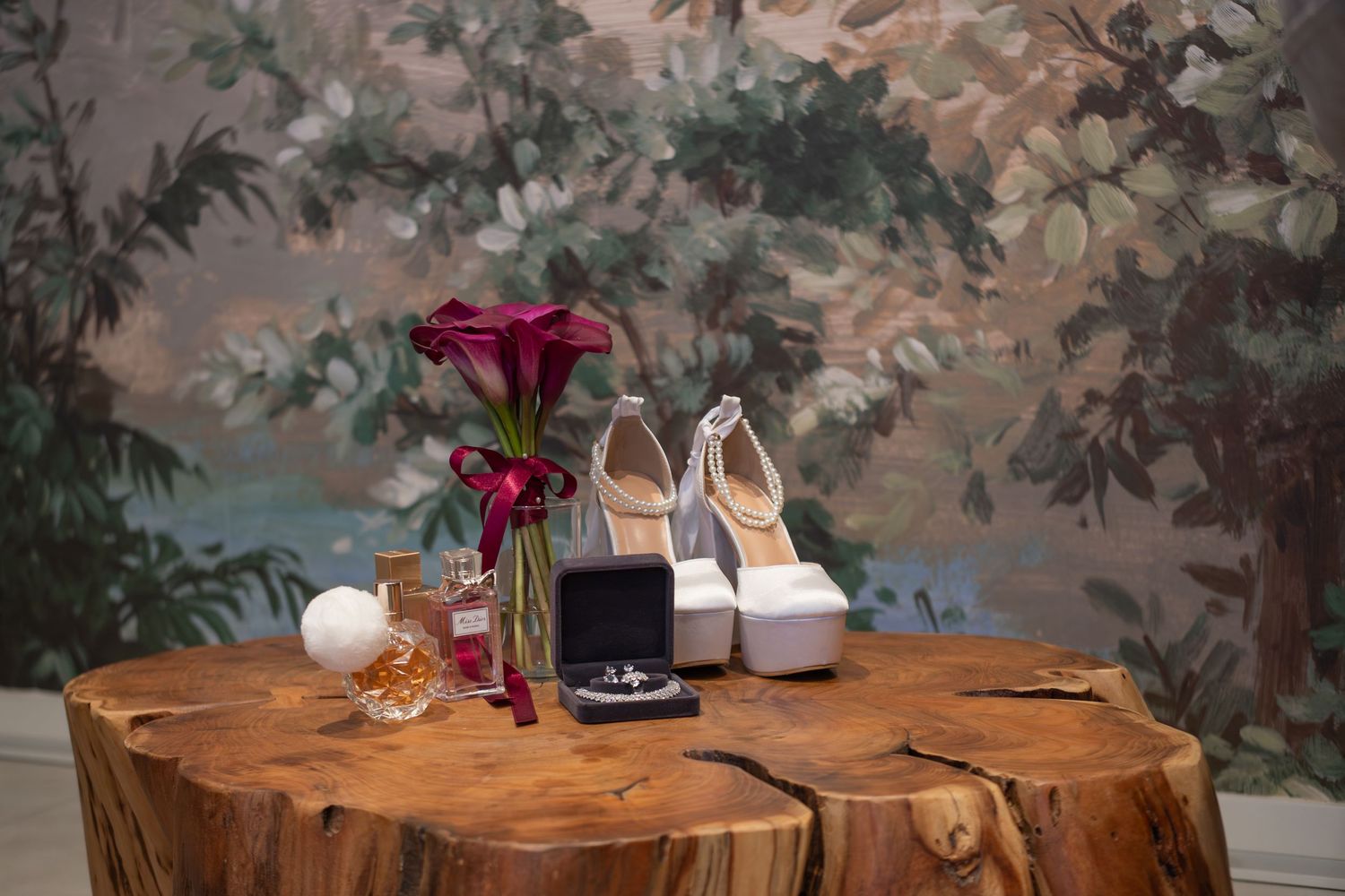 Bridal accessories flat lay featuring white heels, burgundy calla lilies, perfume, and a ring box on a wood stump.