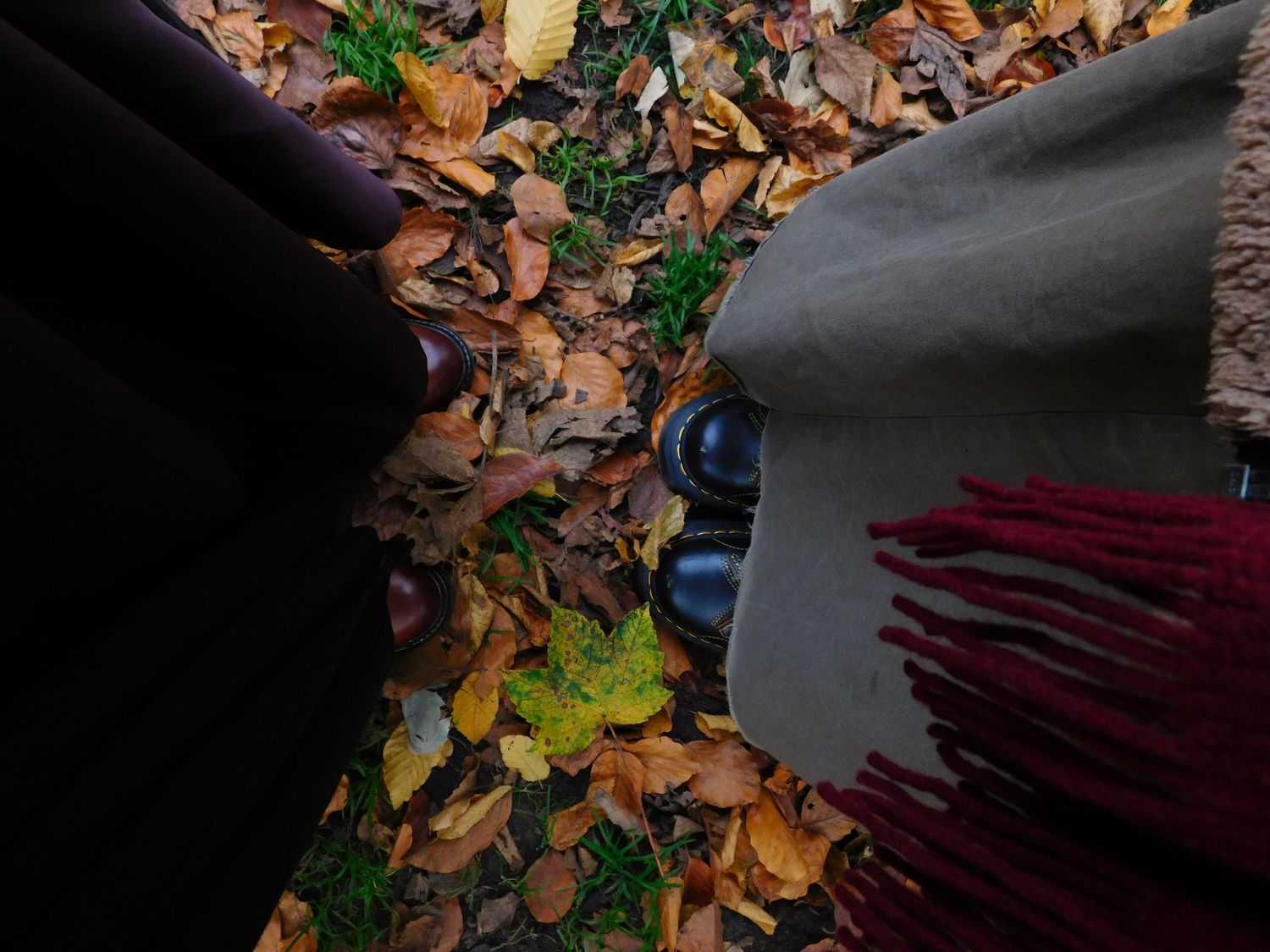 Overhead view of black boots and a grey fringed scarf resting on colorful autumn fallen leaves.