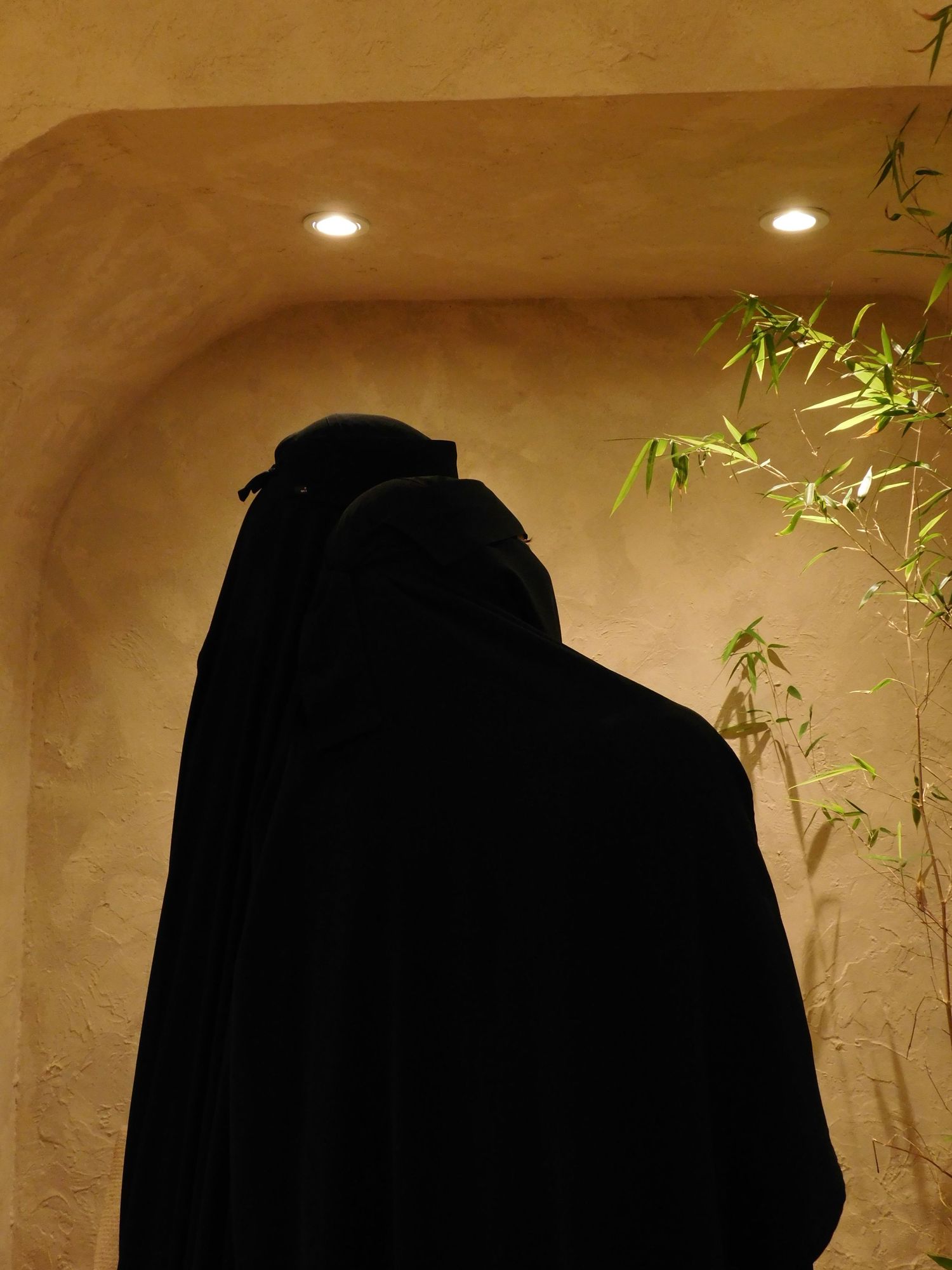 Person wearing traditional black niqab and abaya standing in arched alcove with bamboo plant.