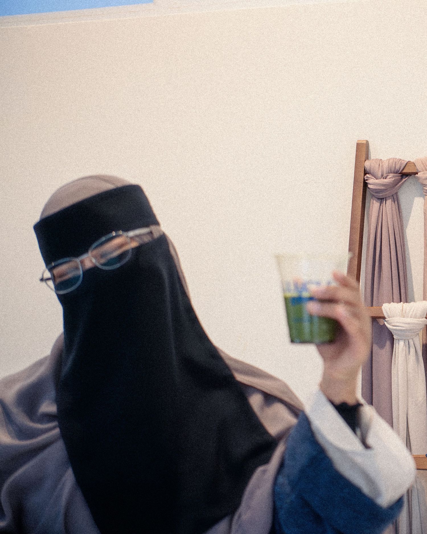 Woman wearing black niqab and glasses holds a cup while standing near a wooden easel.