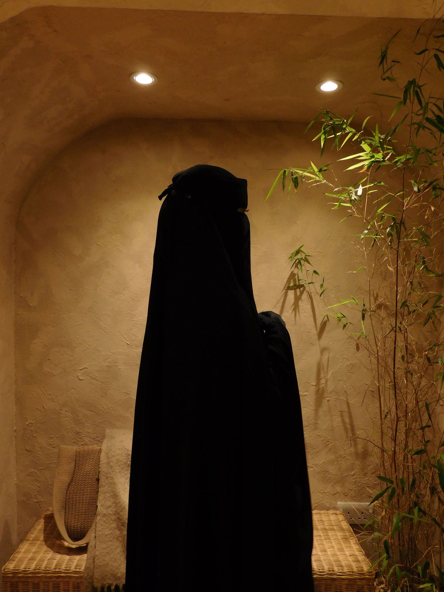 Silhouette of person in black niqab standing in warmly lit arched alcove with bamboo.