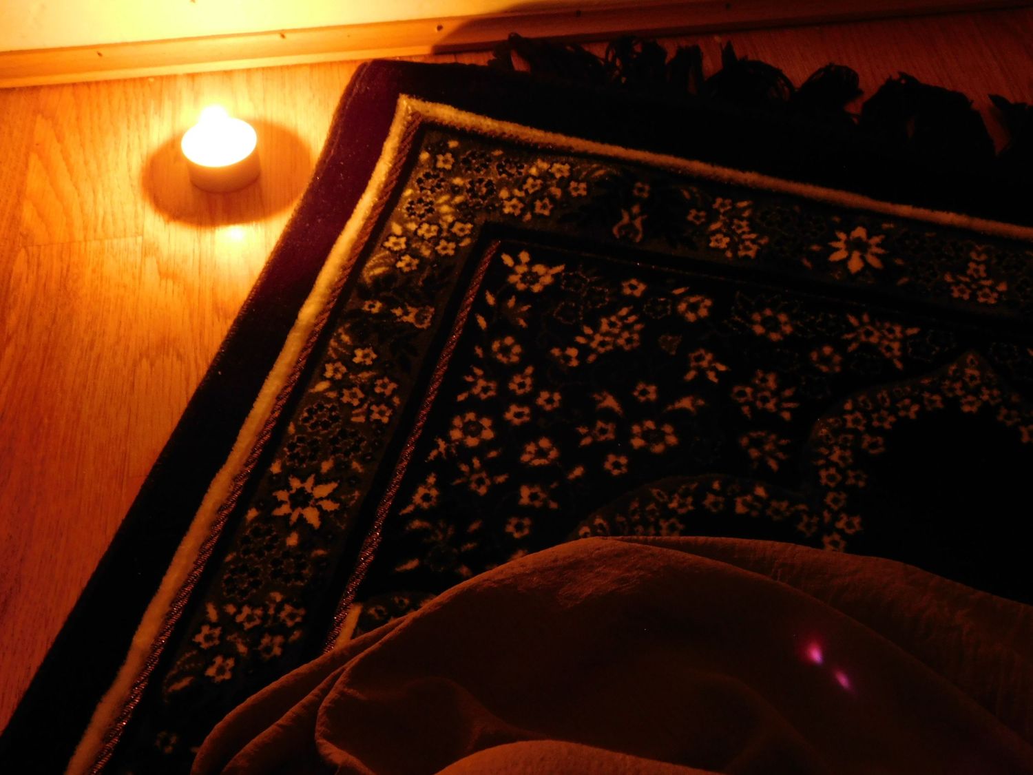 A decorative dark floral rug illuminated by a single glowing tealight candle on a wooden floor.