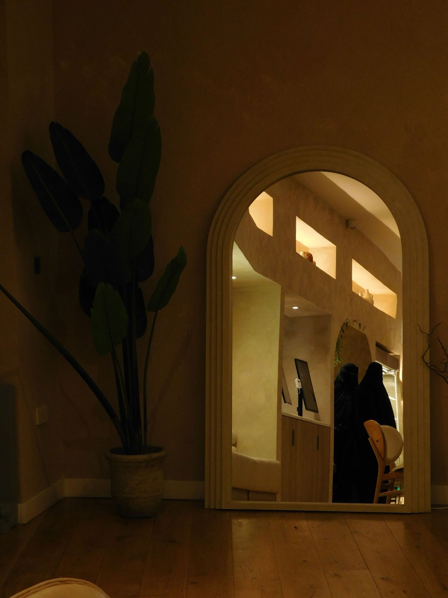 An arched mirror reflects a warmly lit boutique interior with wooden shelving beside a tropical potted plant.