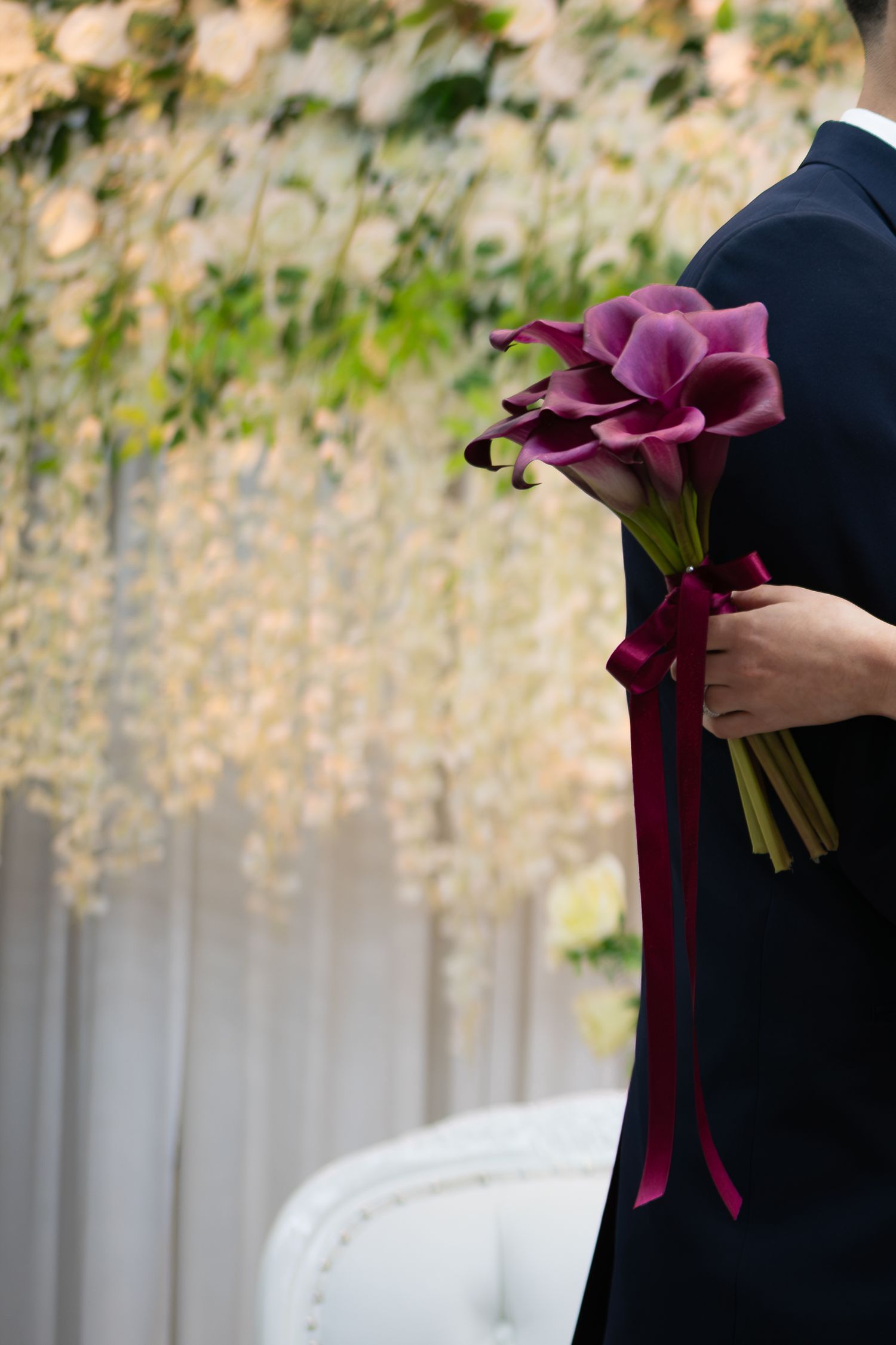 Person holding a bouquet of purple calla lilies tied with a burgundy ribbon at a floral wedding ceremony.