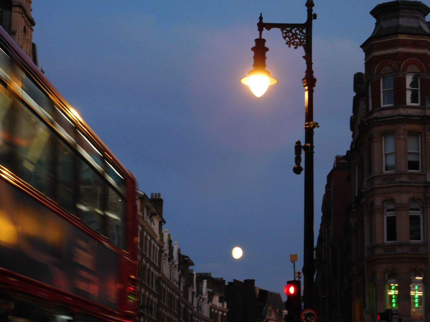 Red double-decker bus passing glowing street lamp on a London street at dusk with full moon.
