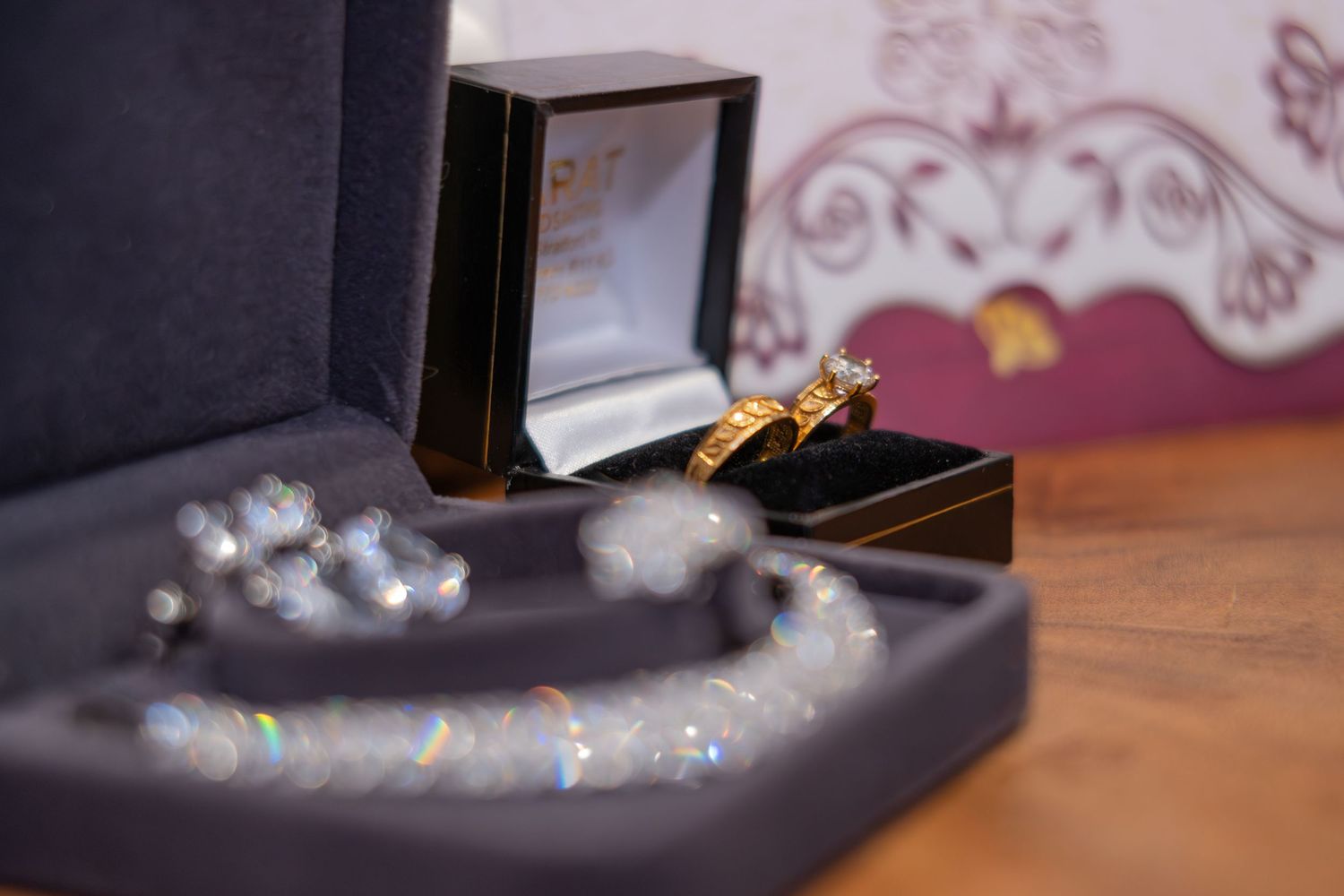 Close-up of a gold engagement ring set in a jewellery box beside a sparkling diamond necklace on a wooden surface.