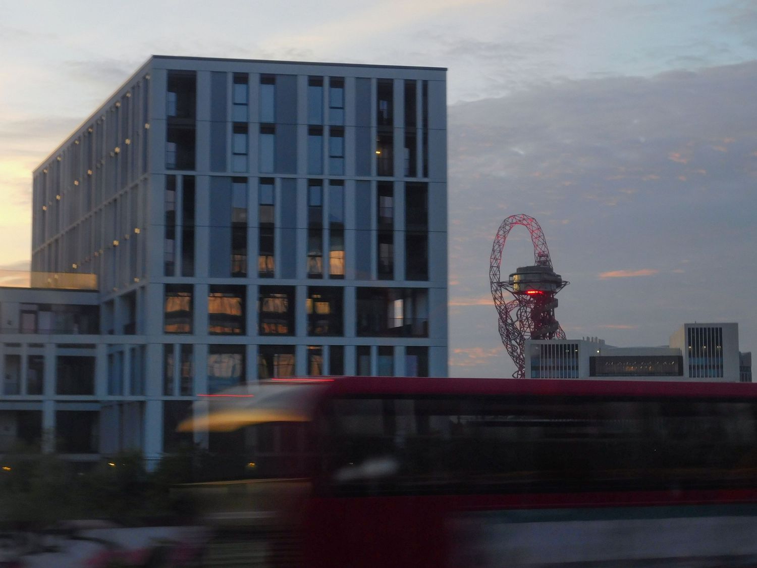 Red London bus passing modern office building with ArcelorMittal Orbit sculpture at sunset.