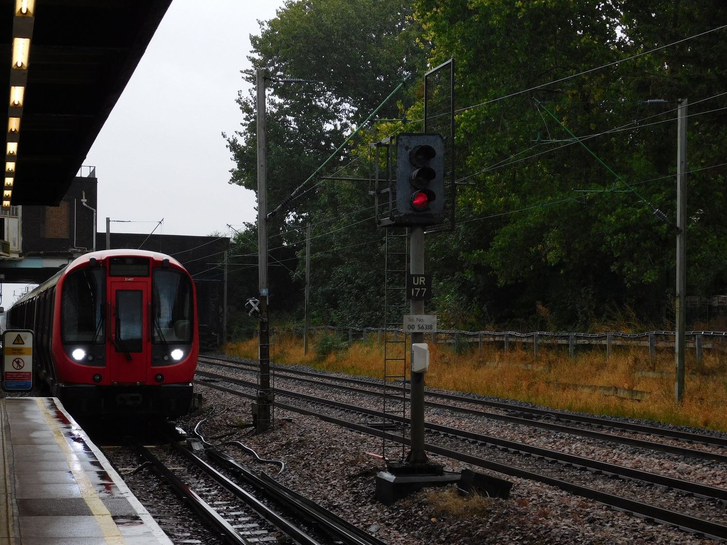 A red London Underground tube train arriving at a suburban station on a rainy overcast day.