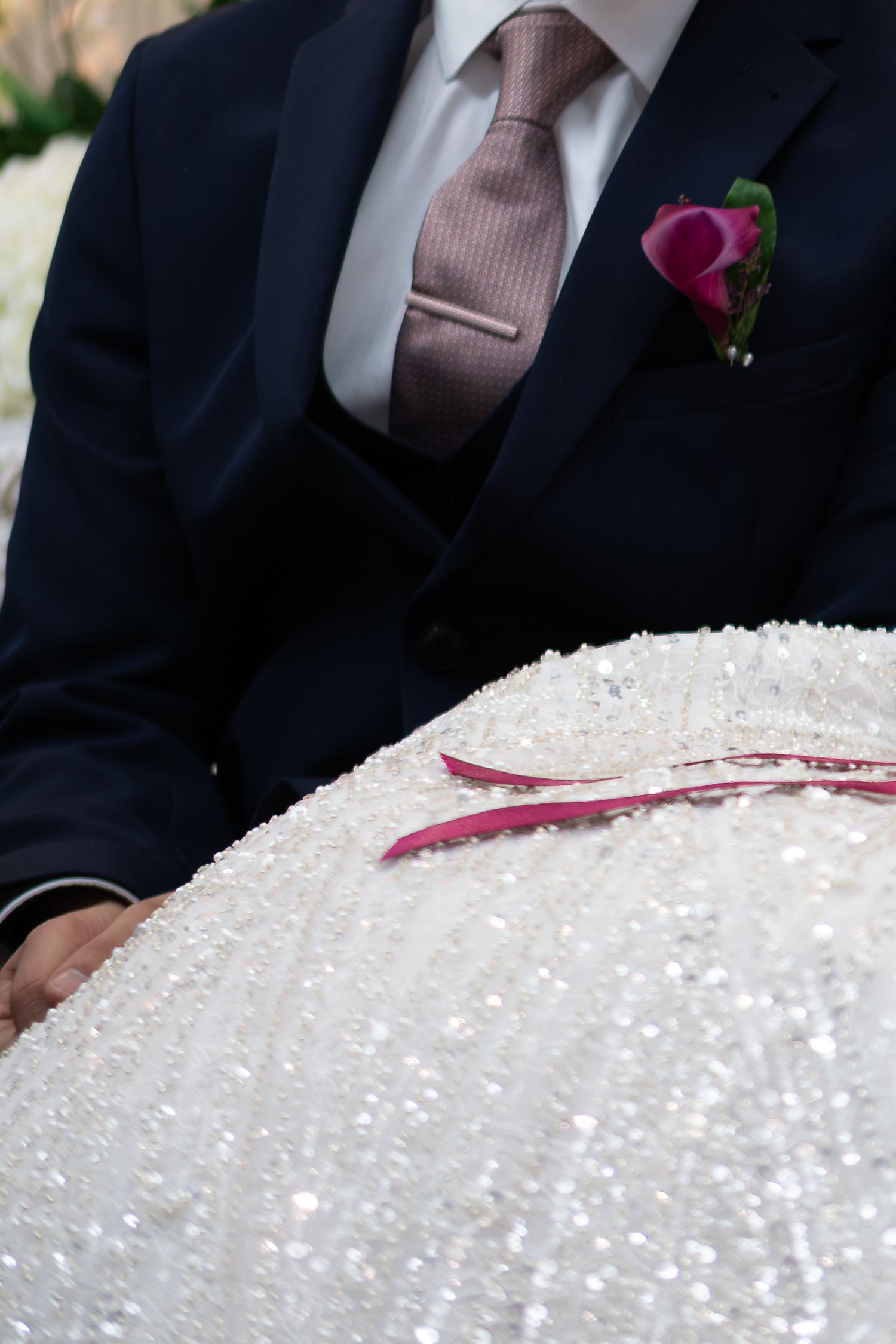 Groom in navy tuxedo with pink boutonniere holding a white beaded wedding item at ceremony.