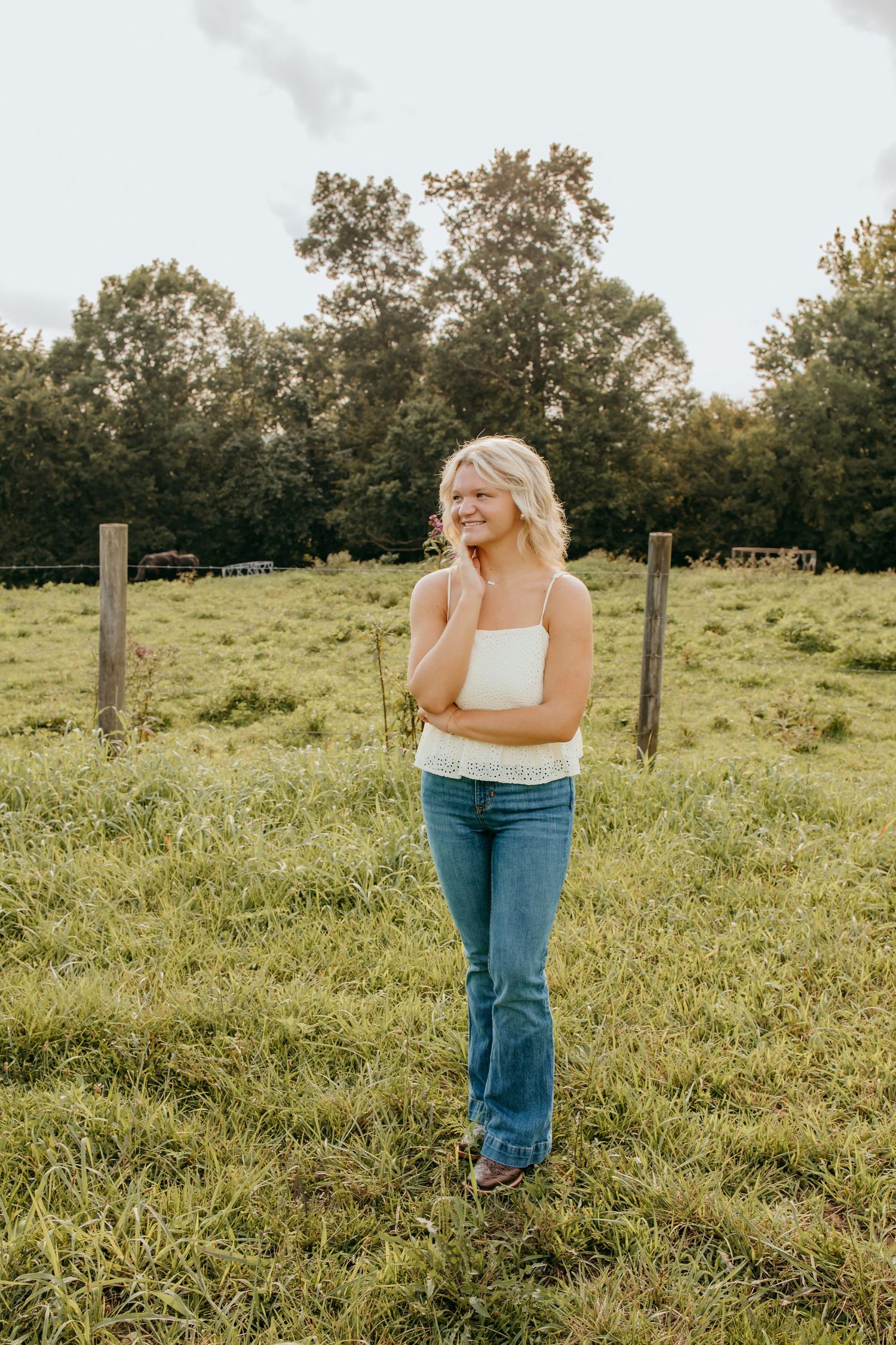 Young blonde woman in white top and jeans smiling in a green pasture field with wooden fence posts and trees.
