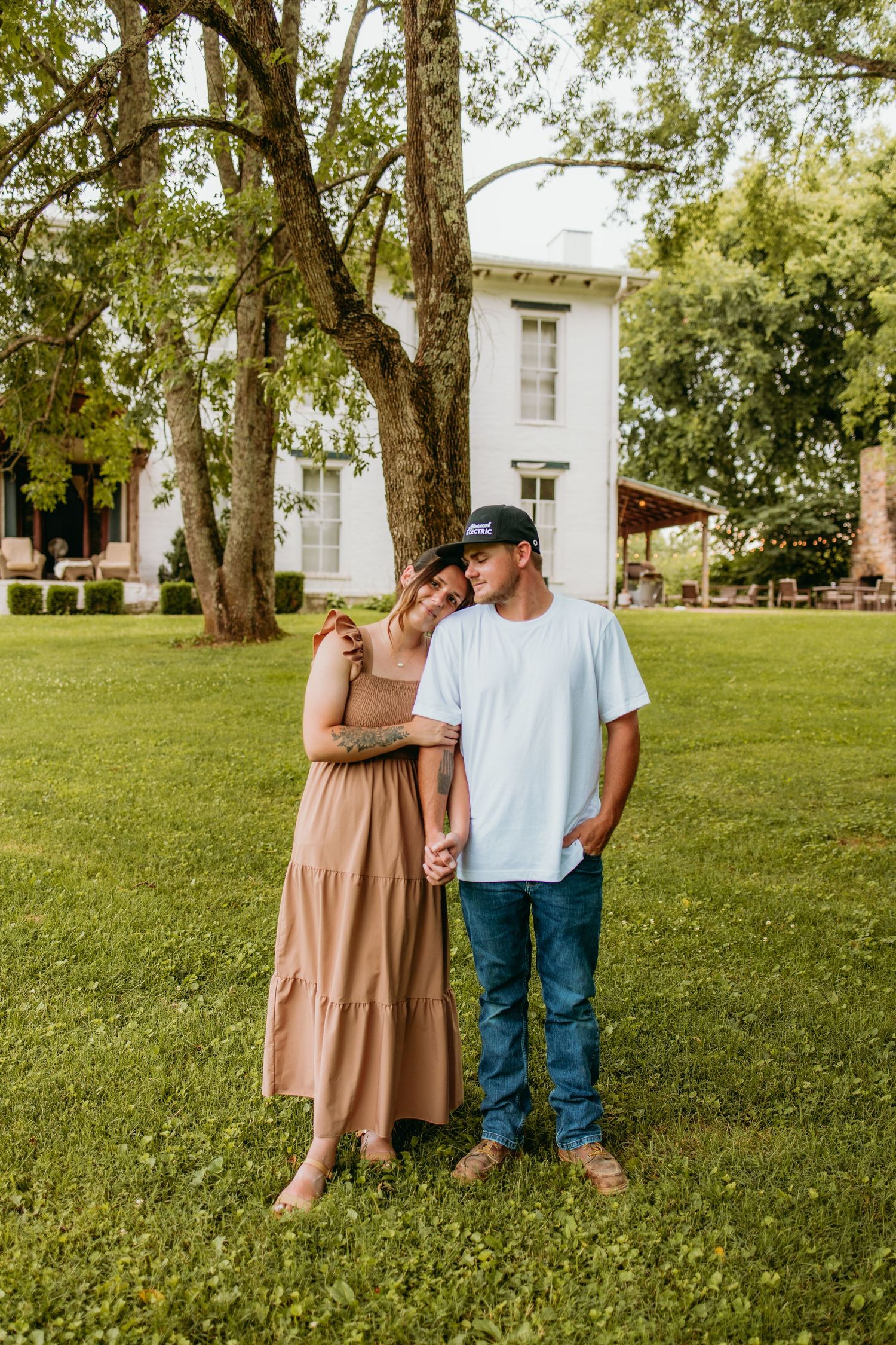 A couple stands hand-in-hand on green grass, woman in tan dress leaning on man in white tee and jeans, white house behind.