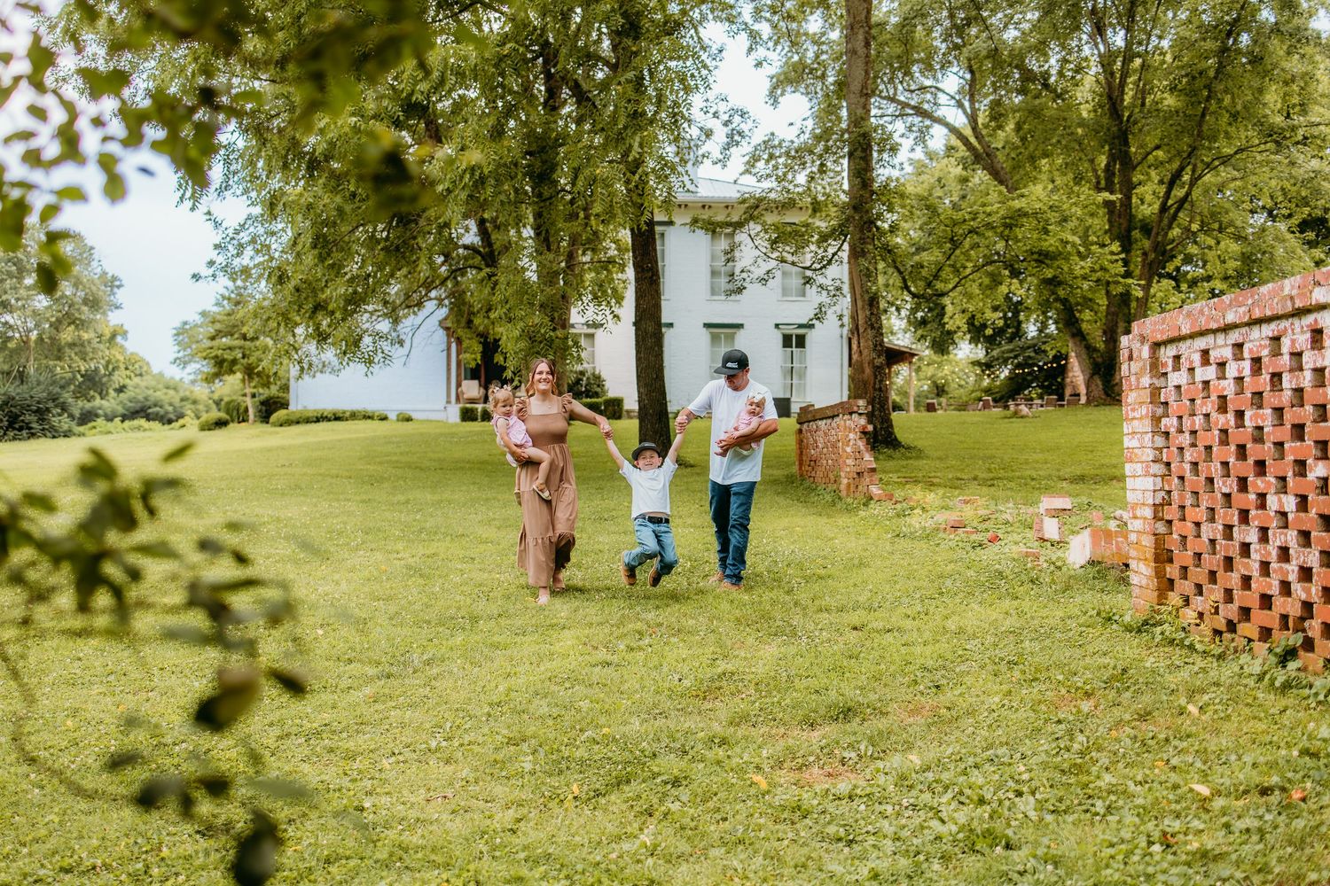 Family of three holding hands and playing in a lush green yard with trees and a white house in the background.