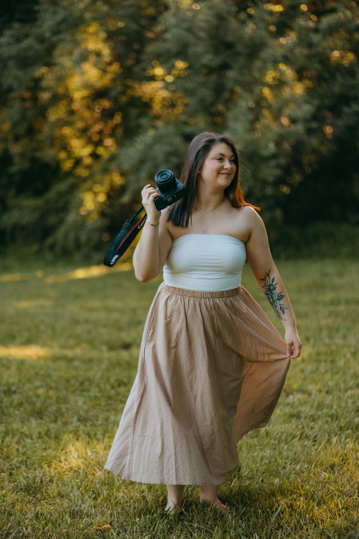 Woman in white tube top and plaid beige maxi skirt holds camera outdoors, showcasing tattoo, smiling in sunlit green park.