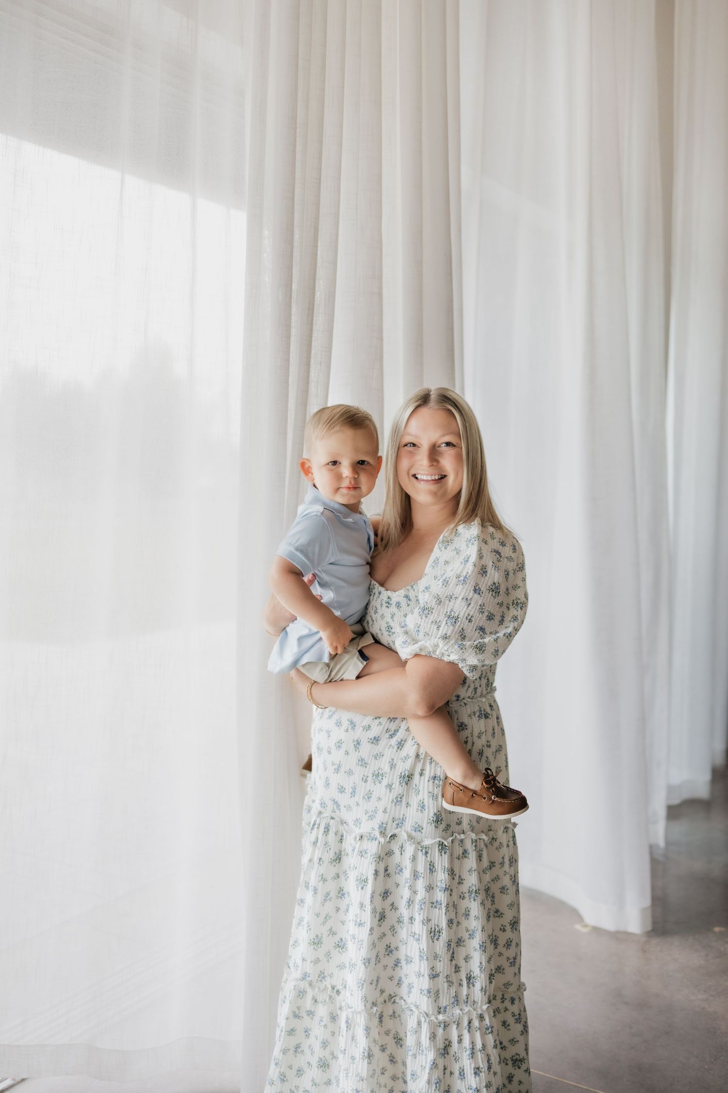 Smiling blonde woman in floral maxi dress holding a toddler boy, posing in front of white sheer curtains.