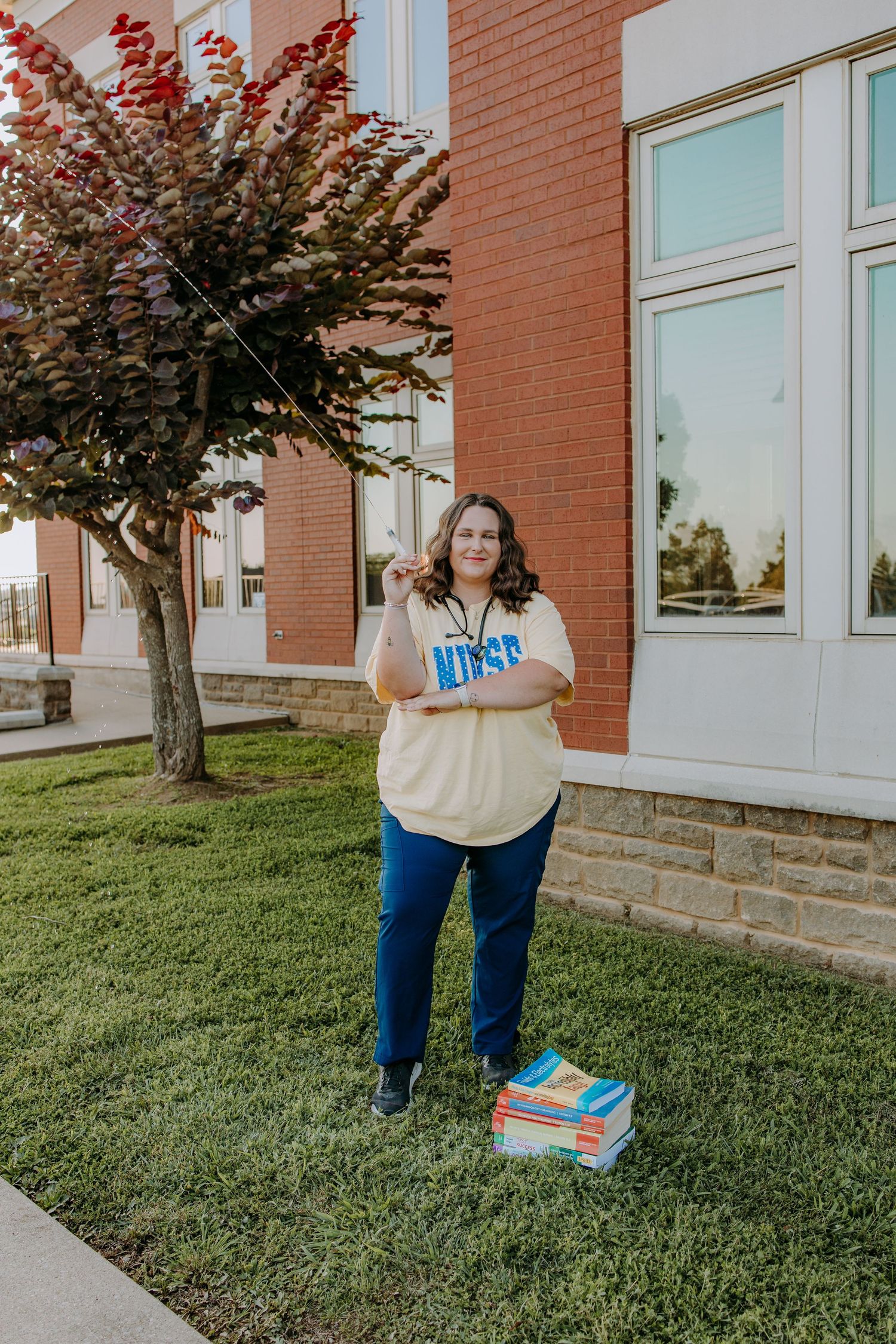 A nursing student stands smiling on a college campus lawn beside stacked textbooks, wearing a yellow 