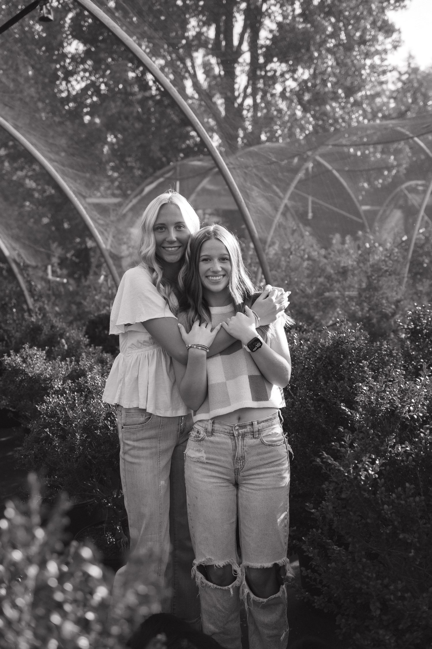 Two smiling seniors pose together in a garden greenhouse setting in black and white photo.