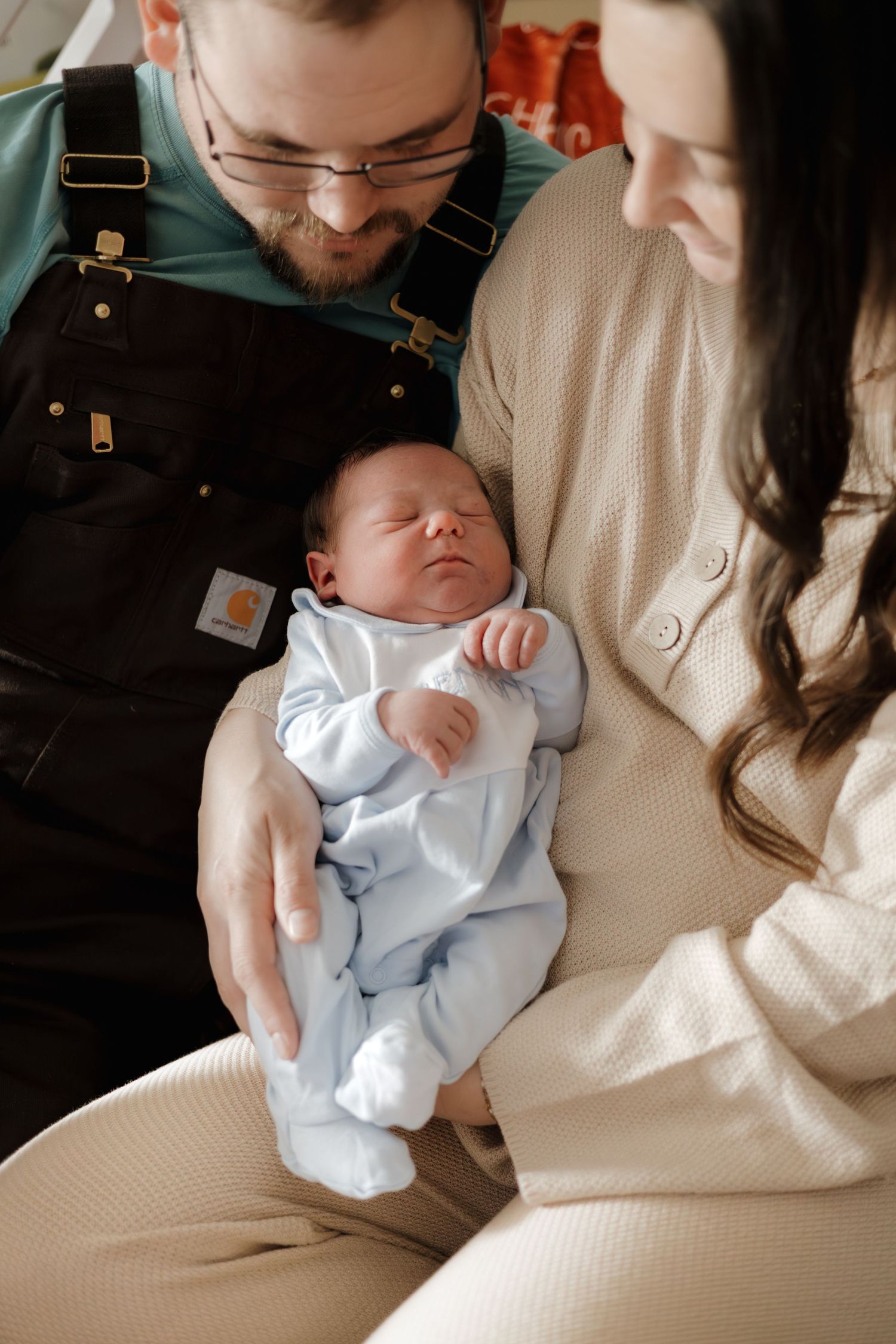 New parents lovingly gaze at their sleeping newborn baby dressed in a light blue onesie.