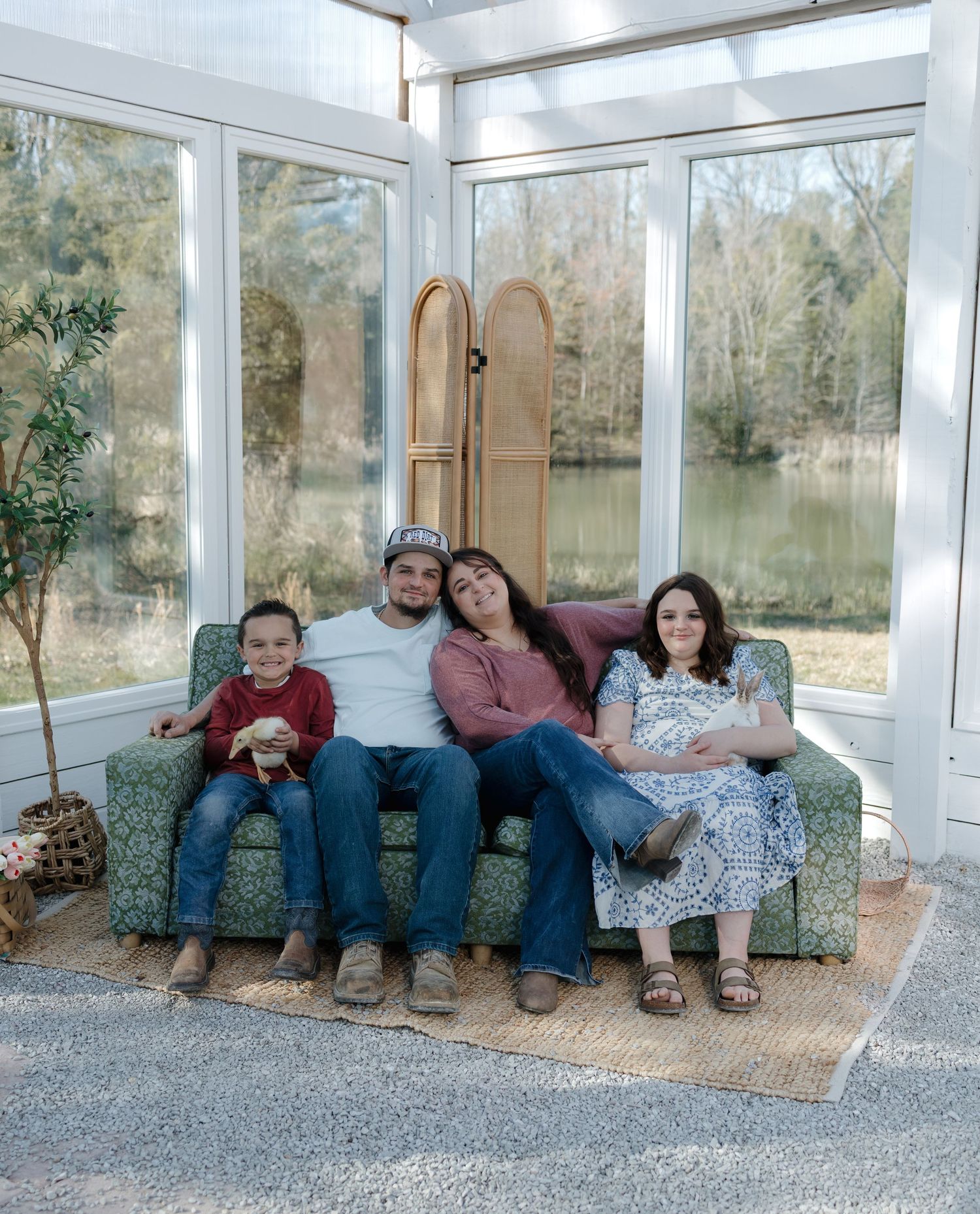 Happy family of four smiling together on green floral sofa in bright sunroom with garden view.