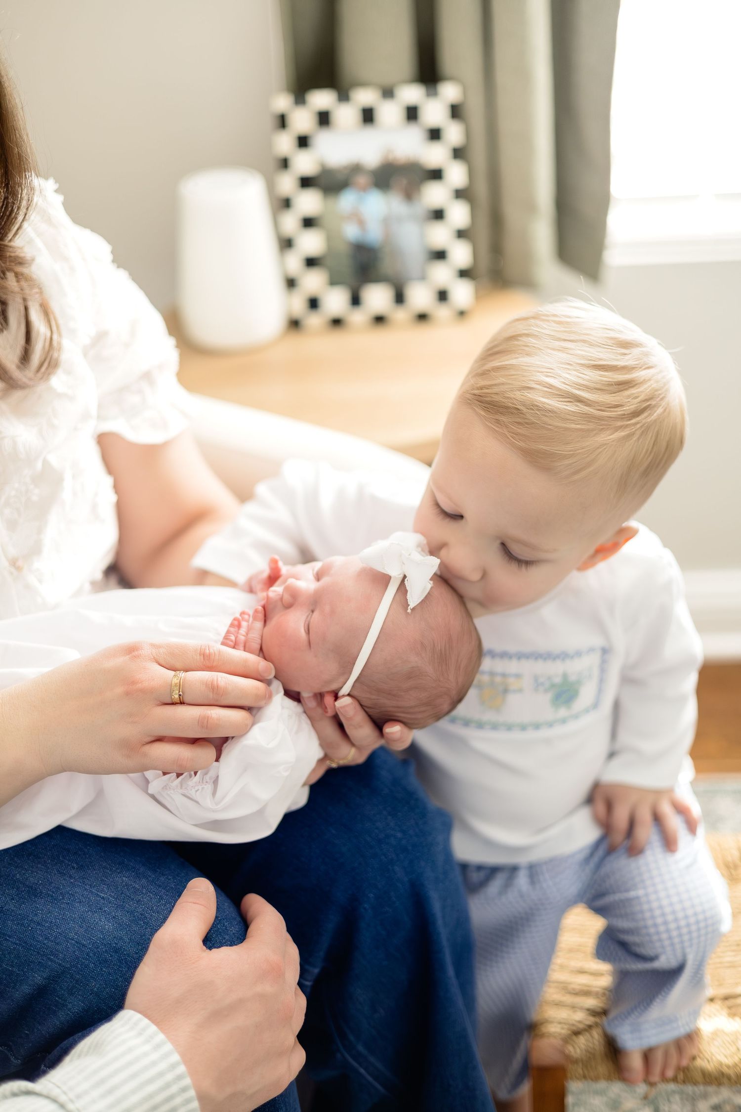 Toddler boy kissing newborn baby sibling held by mother during a sweet family lifestyle newborn photo session.