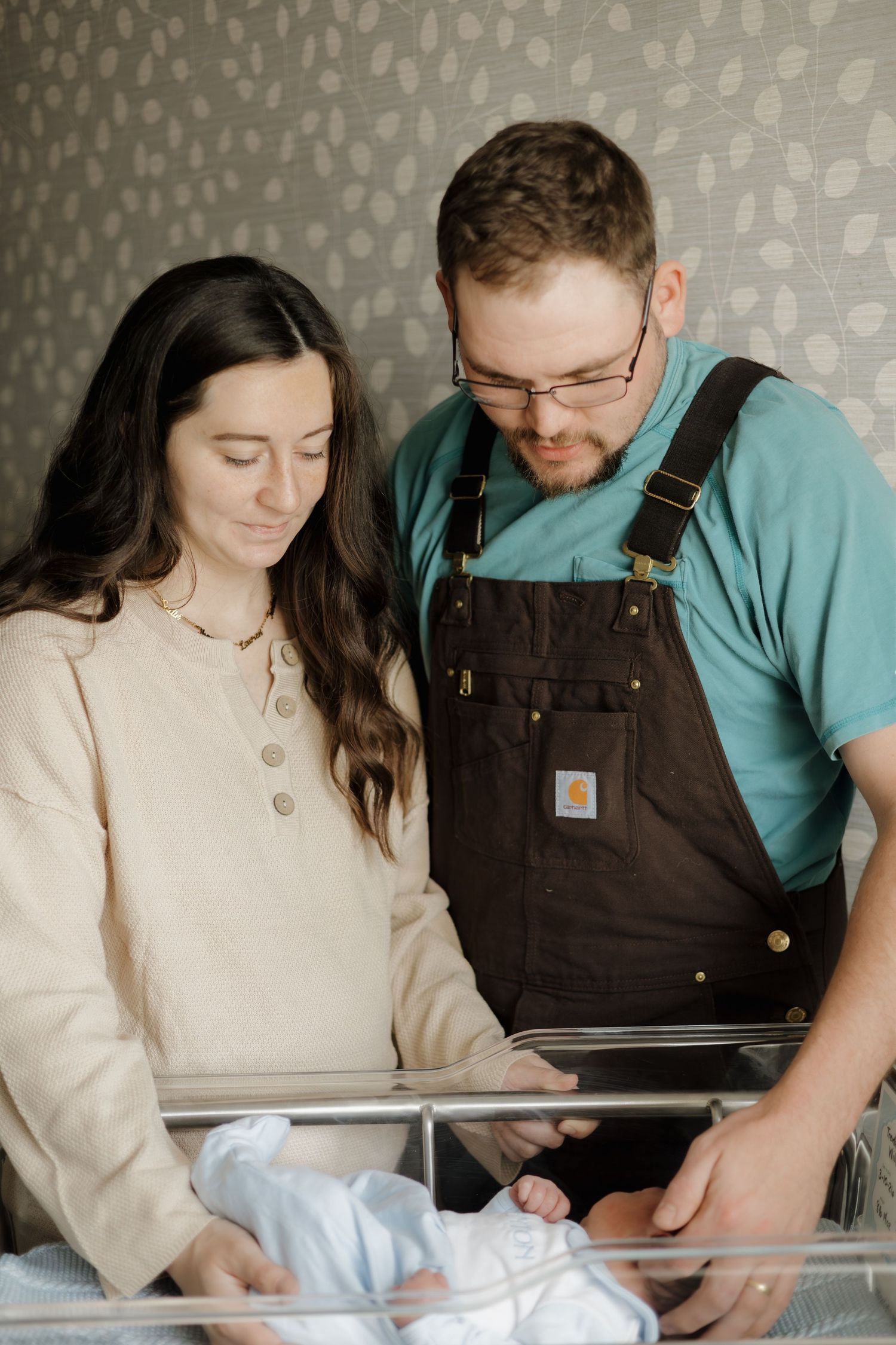 New parents lovingly gaze at their newborn baby in a hospital bassinet, mother in beige top and father in Carhartt overalls.