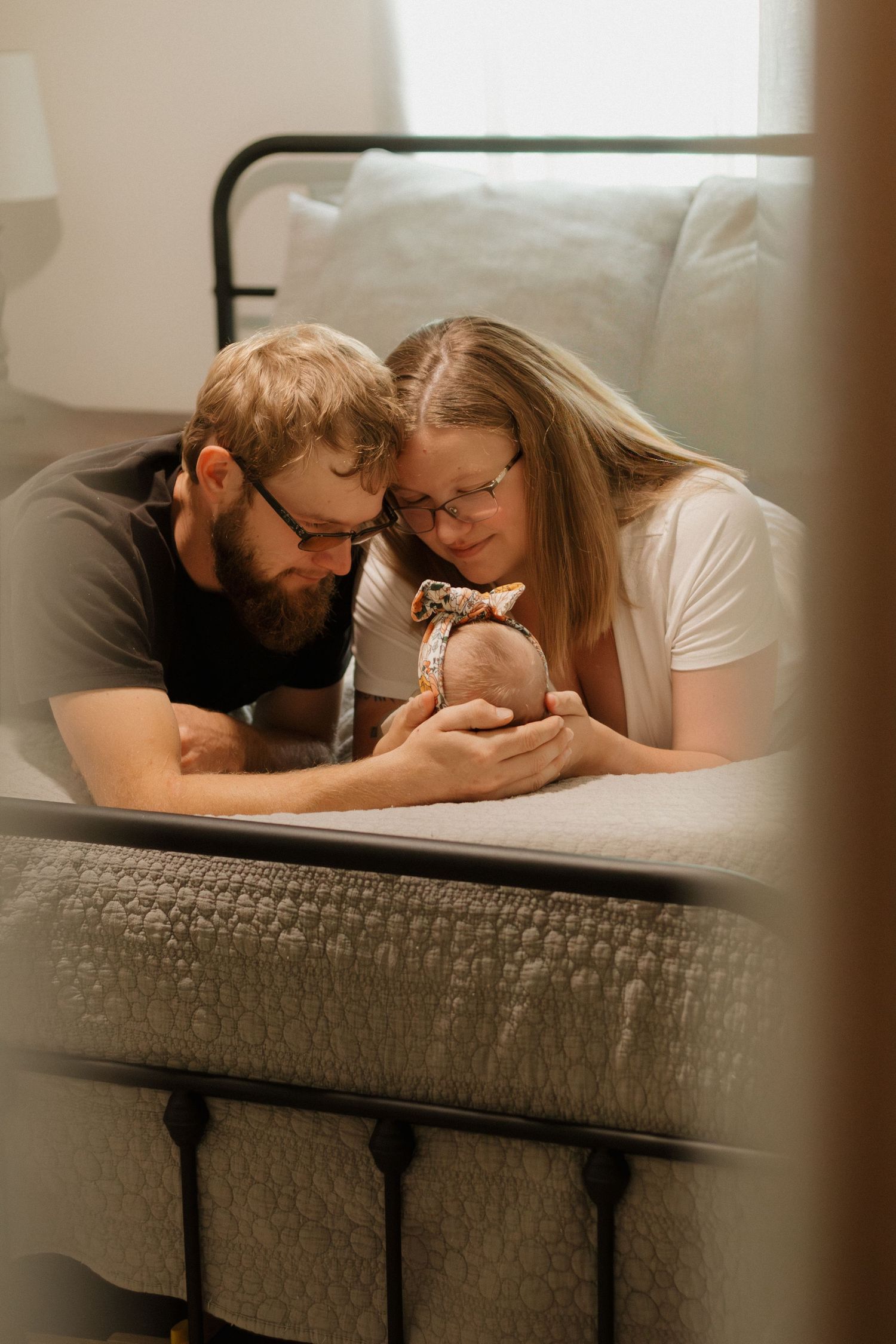 Bearded father and mother with glasses tenderly holding newborn baby together on a bed.