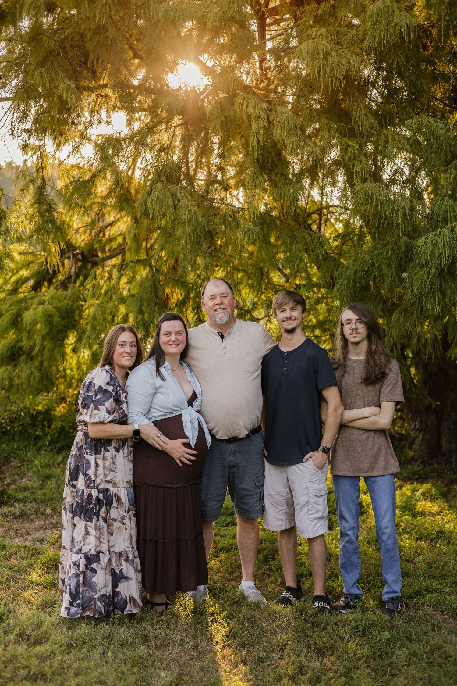 Family of five posing outdoors at golden hour beneath lush green trees, with a pregnant woman in the center.
