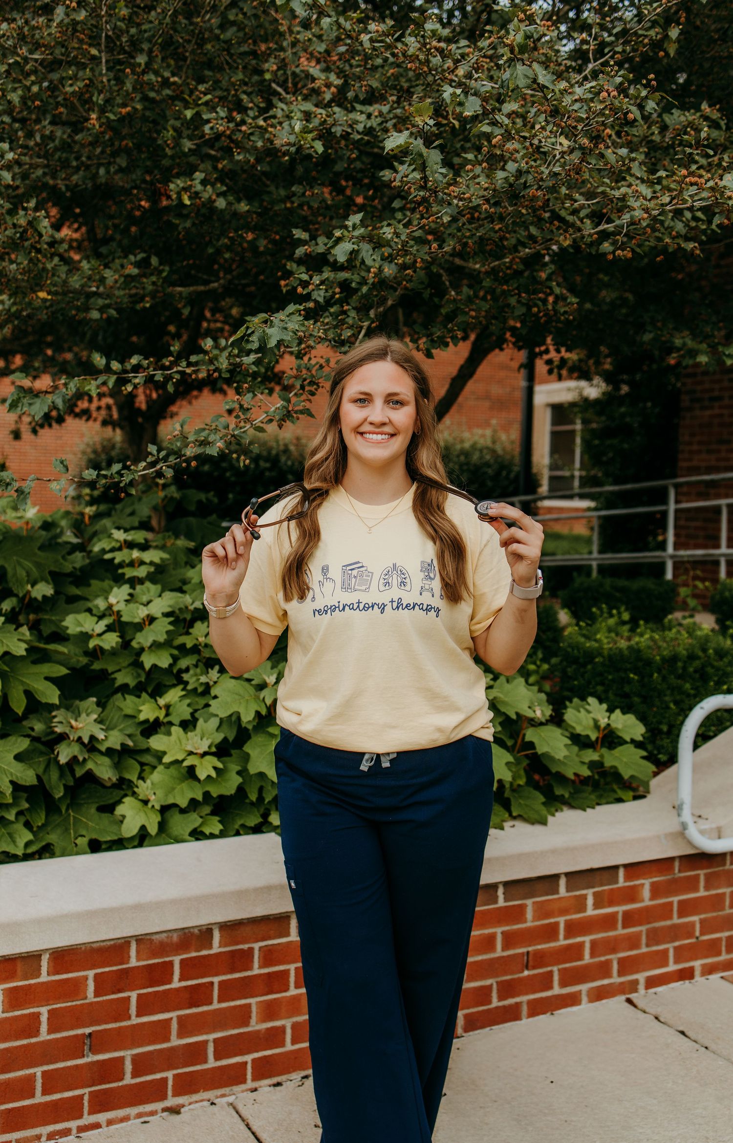 Smiling woman in yellow respiratory therapy t-shirt and navy scrub pants posing outdoors near brick building.