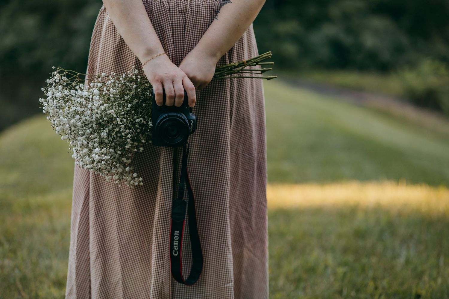 Woman in plaid linen maxi skirt holding a Canon camera and white baby's breath flowers outdoors.