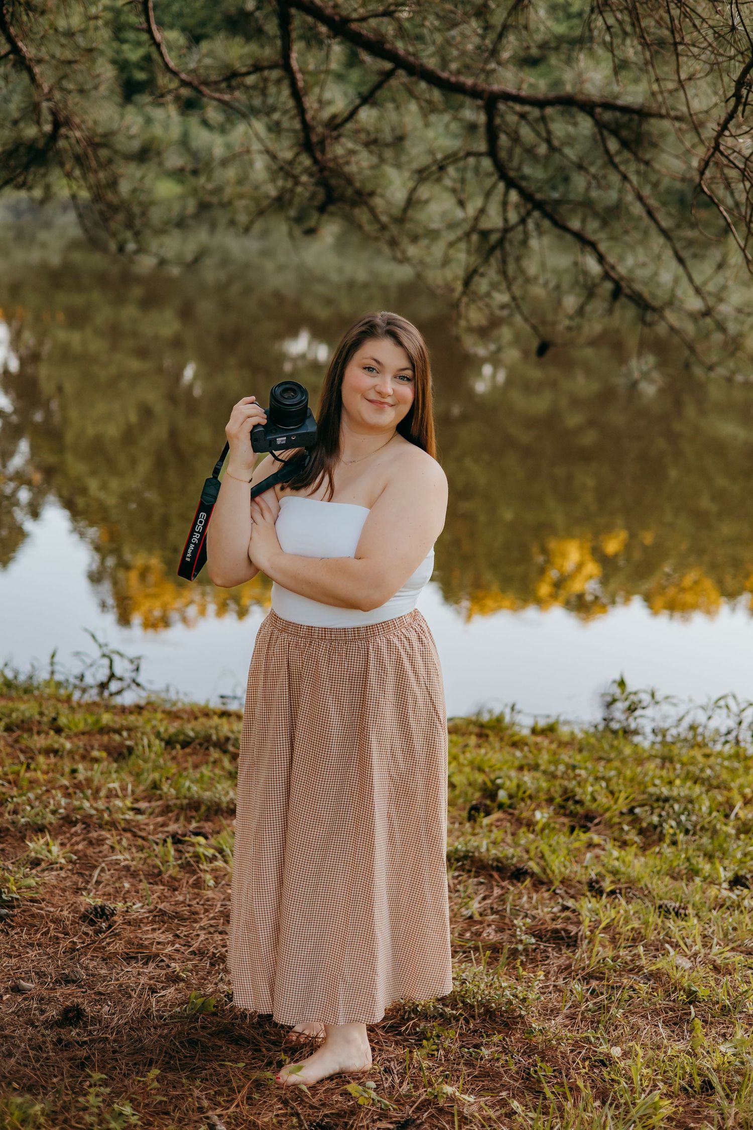 Woman in white top and plaid skirt holds camera near reflective lake surrounded by trees.