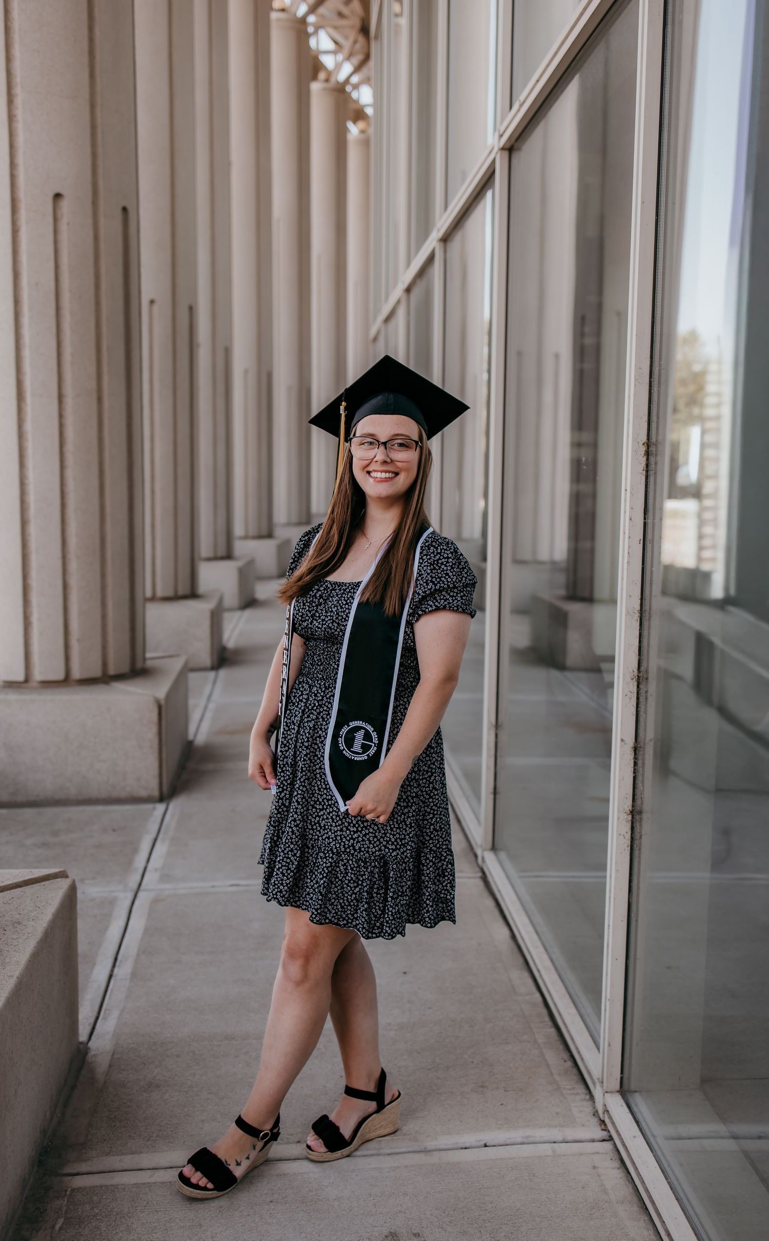 A smiling graduate poses in a black floral dress and cap with a stole outside a modern building with tall columns.
