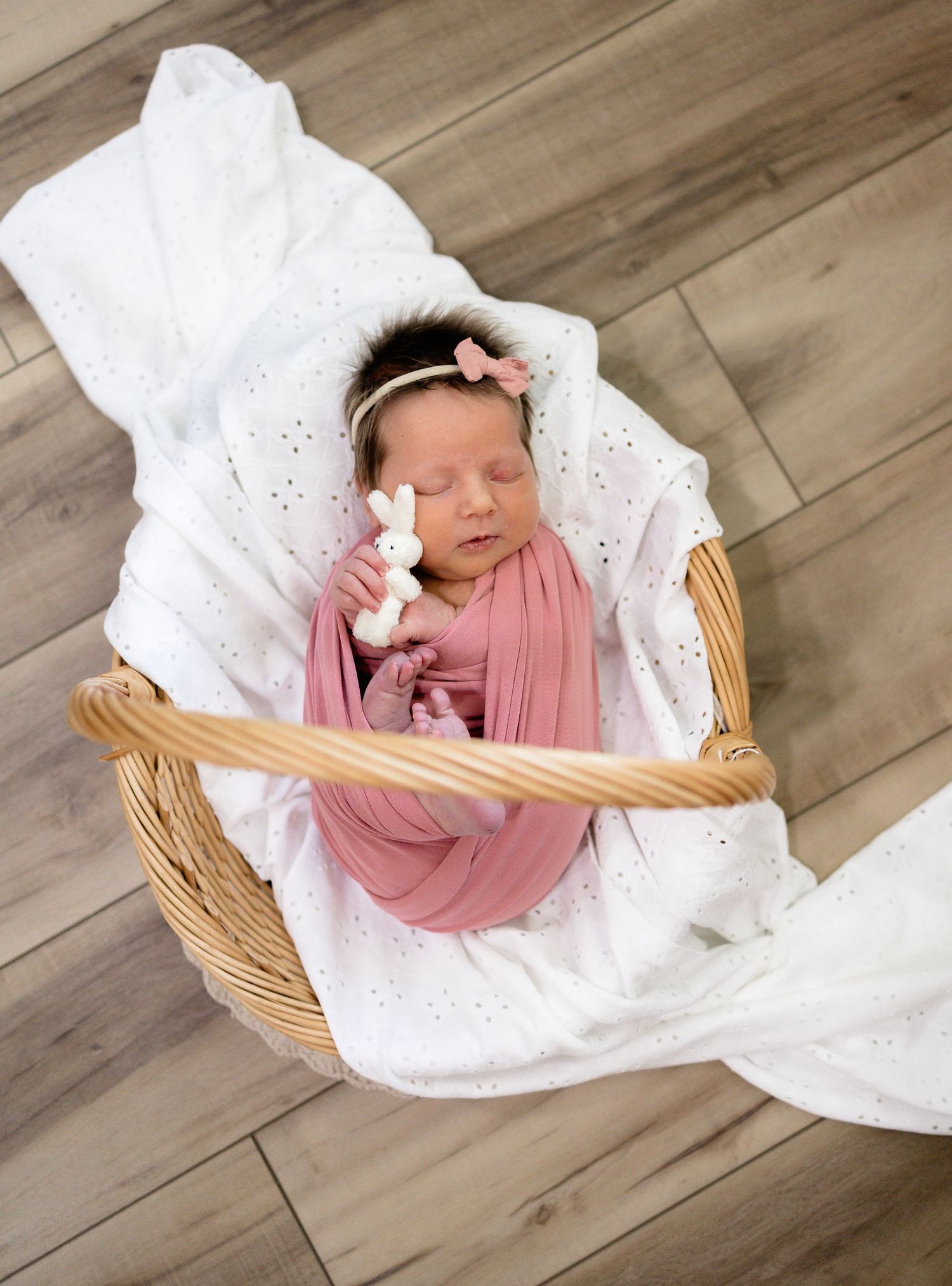 Newborn baby swaddled in pink wrap, sleeping in wicker basket with white blanket and tiny bunny toy, wearing pink bow headband.