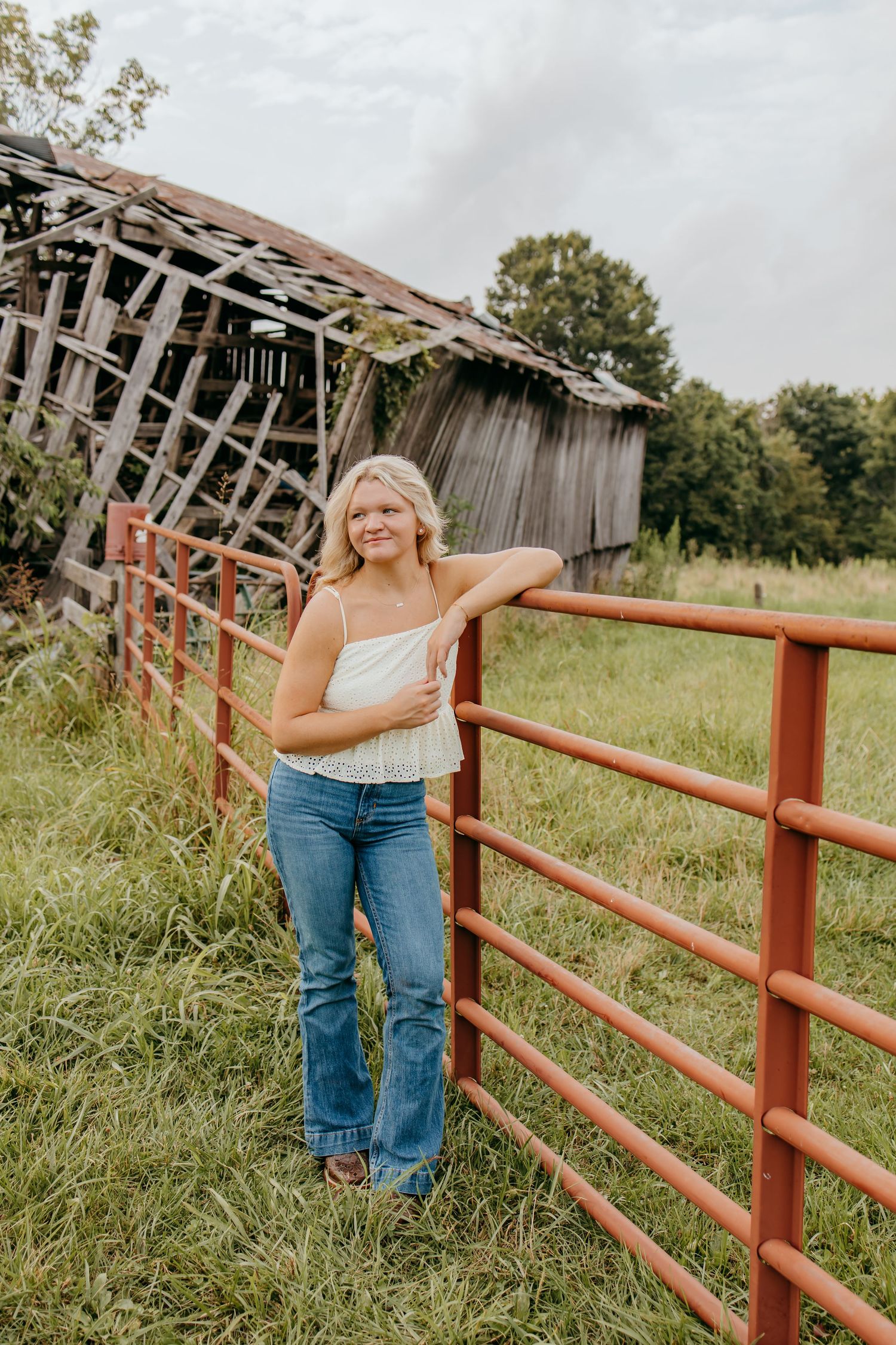 Young blonde woman in white top and jeans leaning on red farm gate with rustic barn in background.
