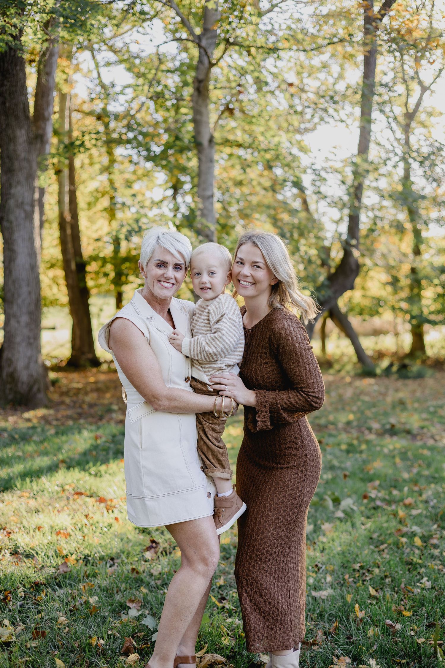 Two blonde women and a toddler pose outdoors in fall foliage, wearing neutral tones in a sunlit park setting.