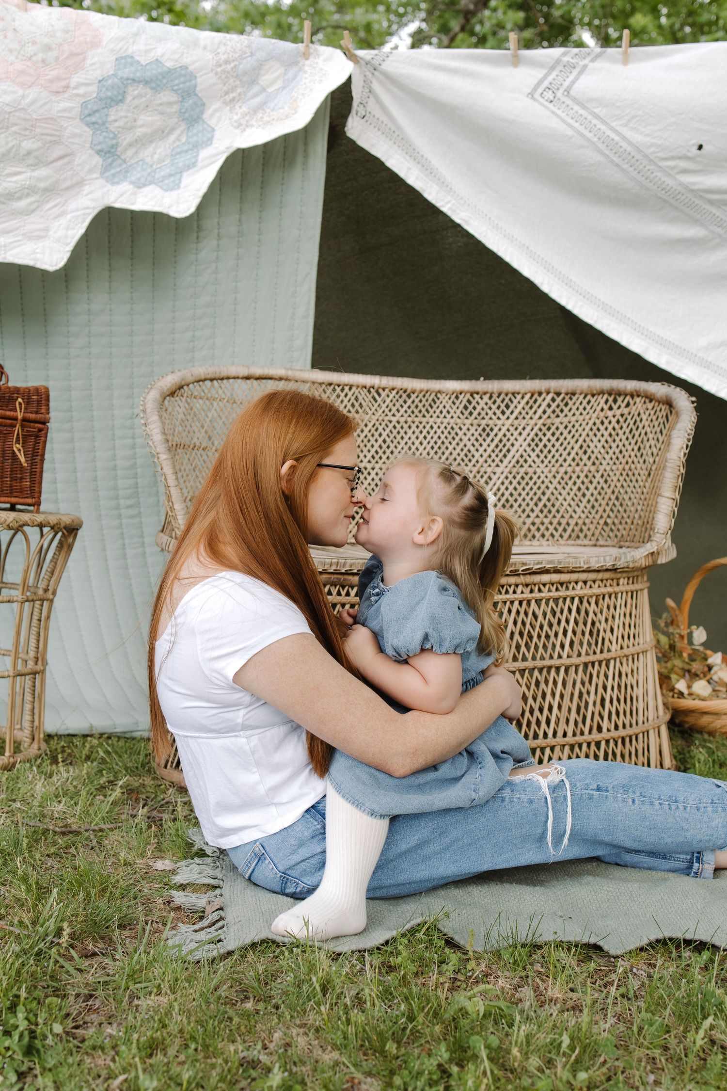 A red-haired mother in white tee and jeans shares an Eskimo kiss with her toddler daughter outdoors near a boho tent.