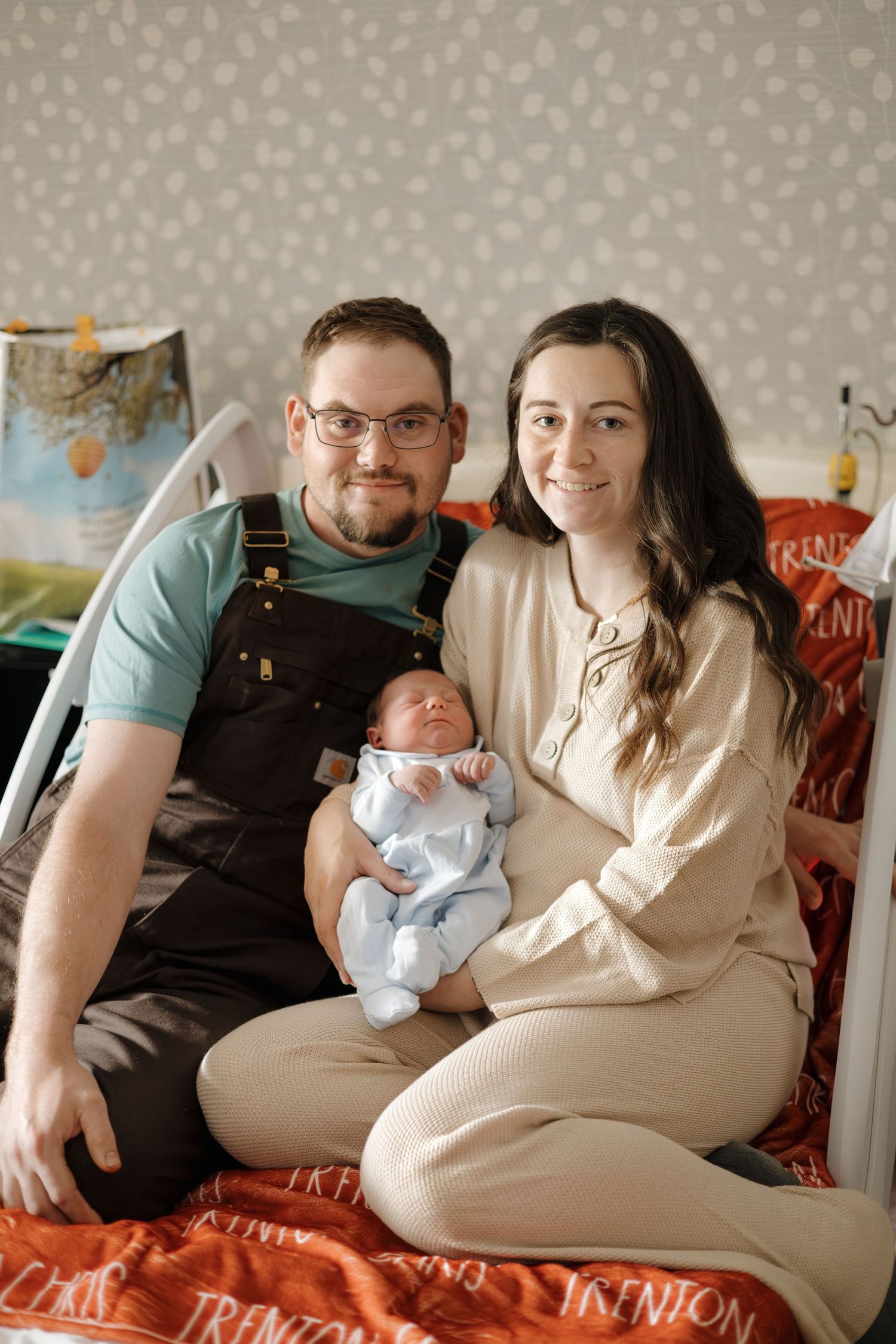 A couple sits on a bed smiling, holding their newborn baby dressed in a light blue outfit in a decorated nursery.
