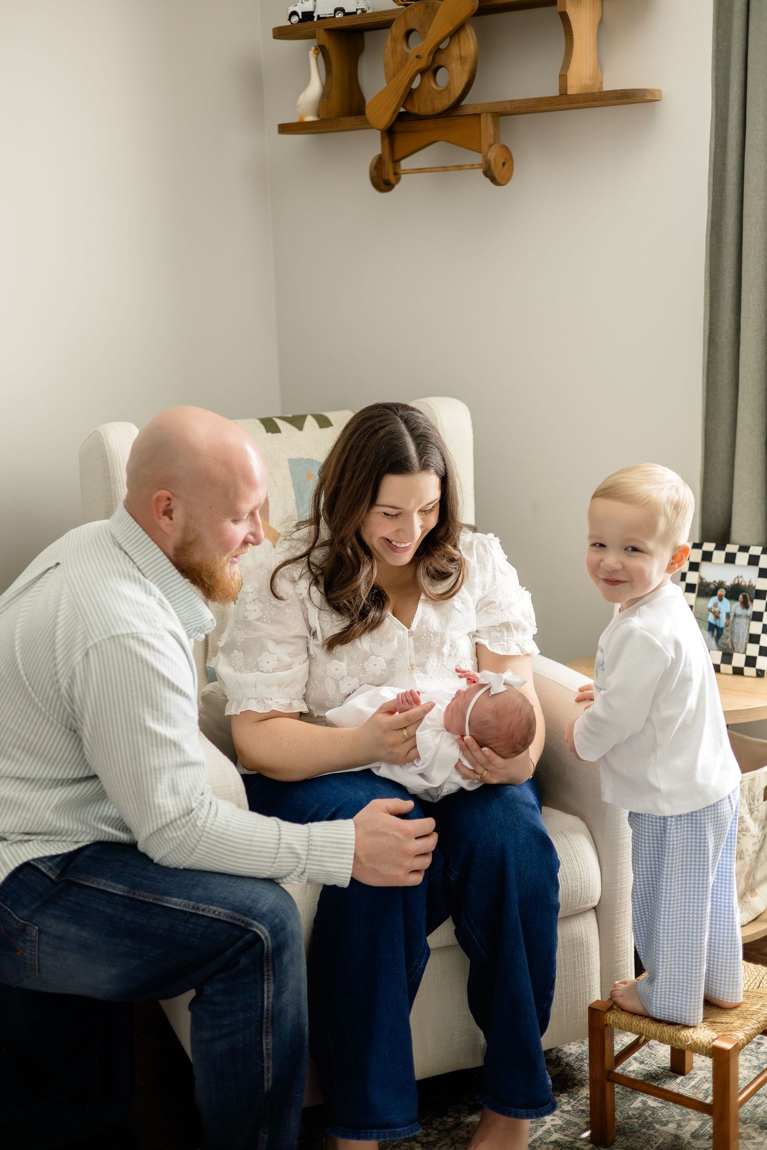 A happy family of four admires their newborn baby in a cozy nursery with a wooden airplane shelf decor.