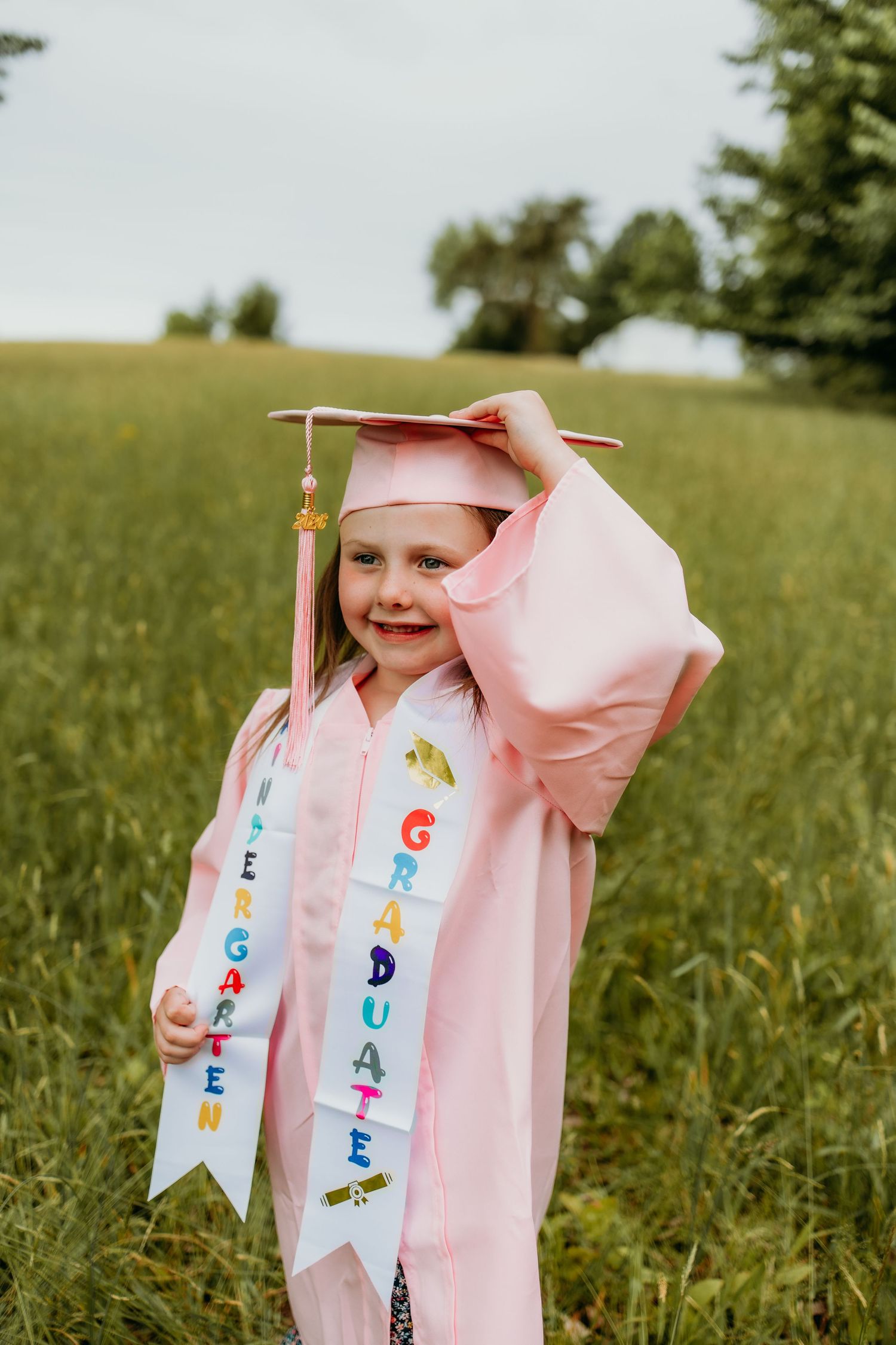 Young girl in pink cap and gown holding a kindergarten graduate sash, smiling outdoors in a green field.