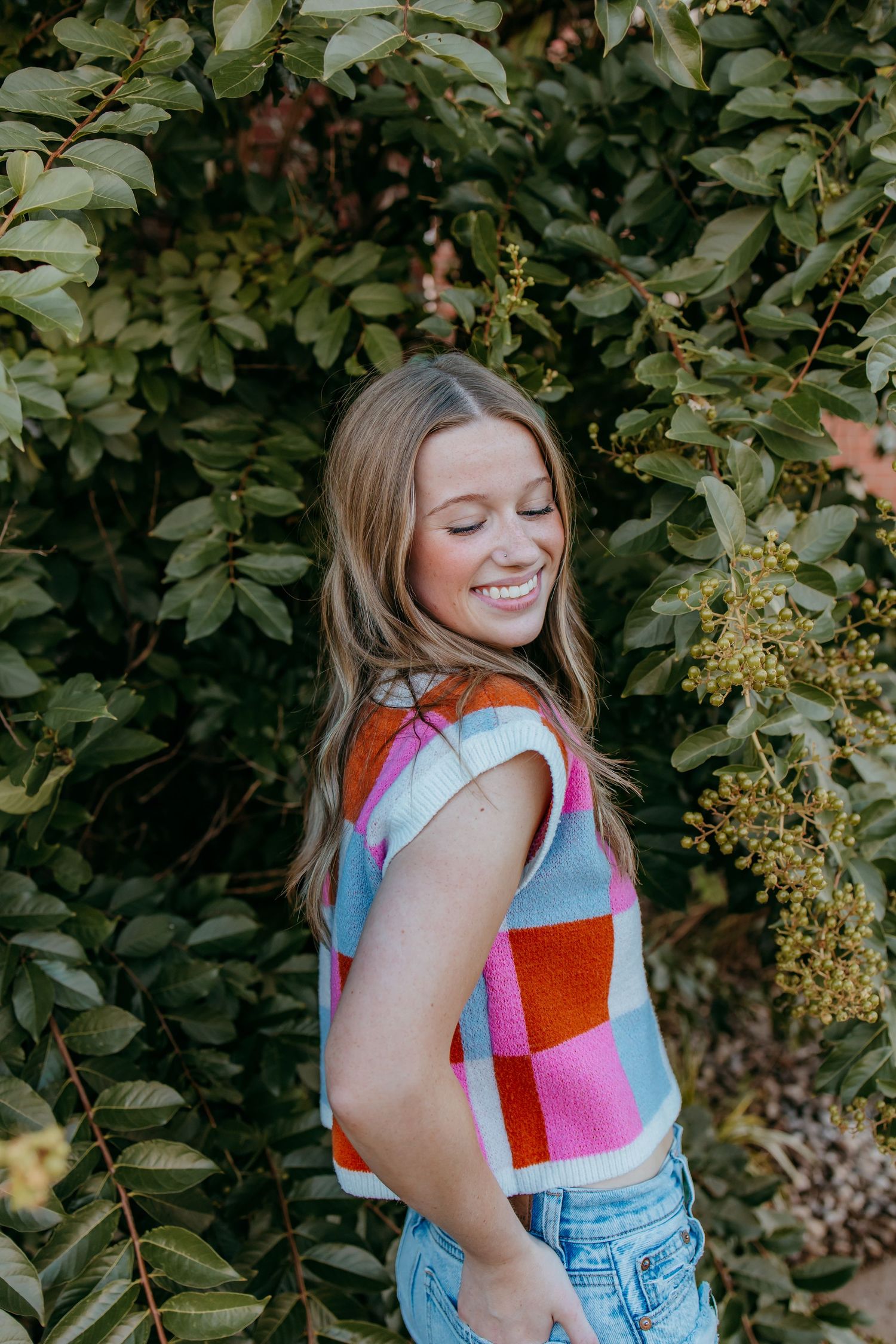 Smiling young woman in colorful checkered crop top and jeans posing outdoors among lush green foliage.