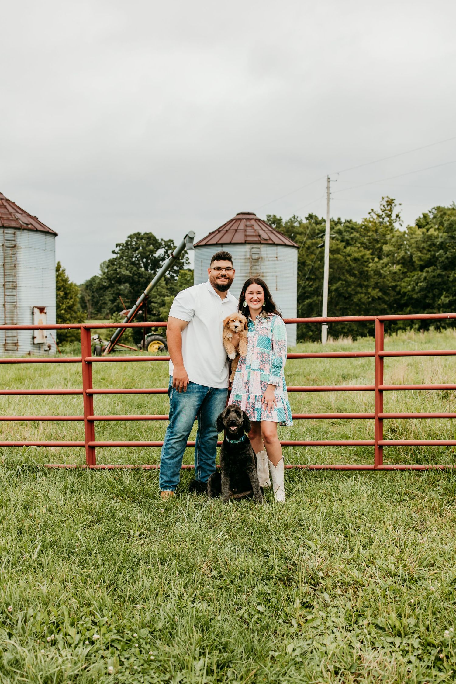 Couple smiling with two dogs by a red farm gate, grain silos and green fields in the background.