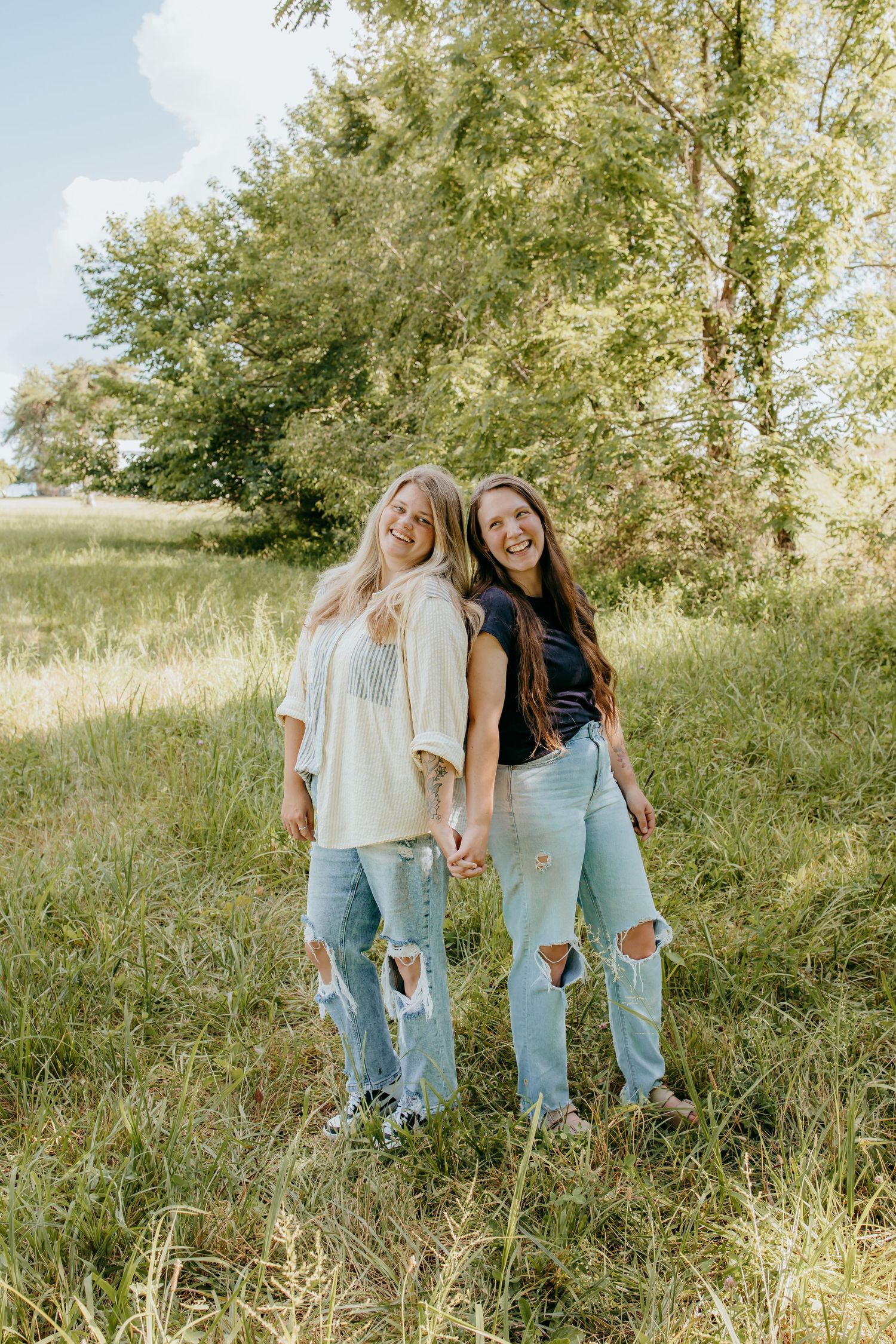Two smiling young women in ripped jeans hold hands in a lush green field on a sunny day.