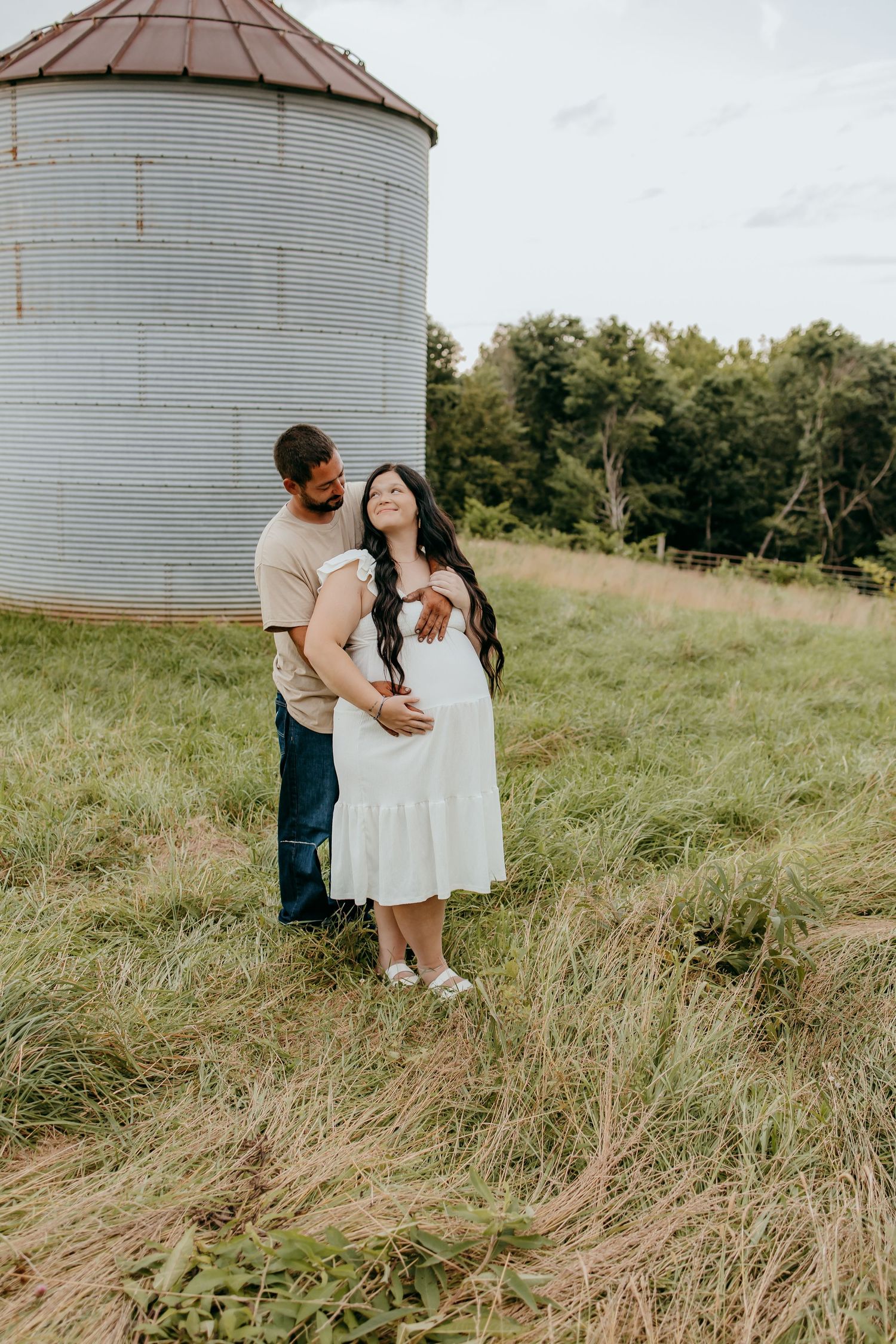 Pregnant woman in white dress with partner embracing outdoors near grain silo on lush green farm.