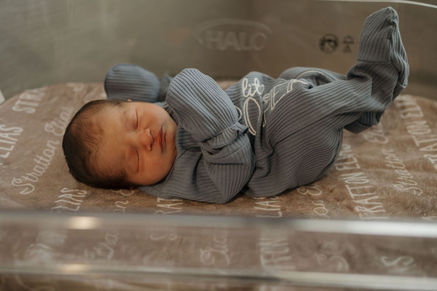 Newborn baby sleeping peacefully in a hospital bassinet wearing a blue outfit on a patterned blanket.