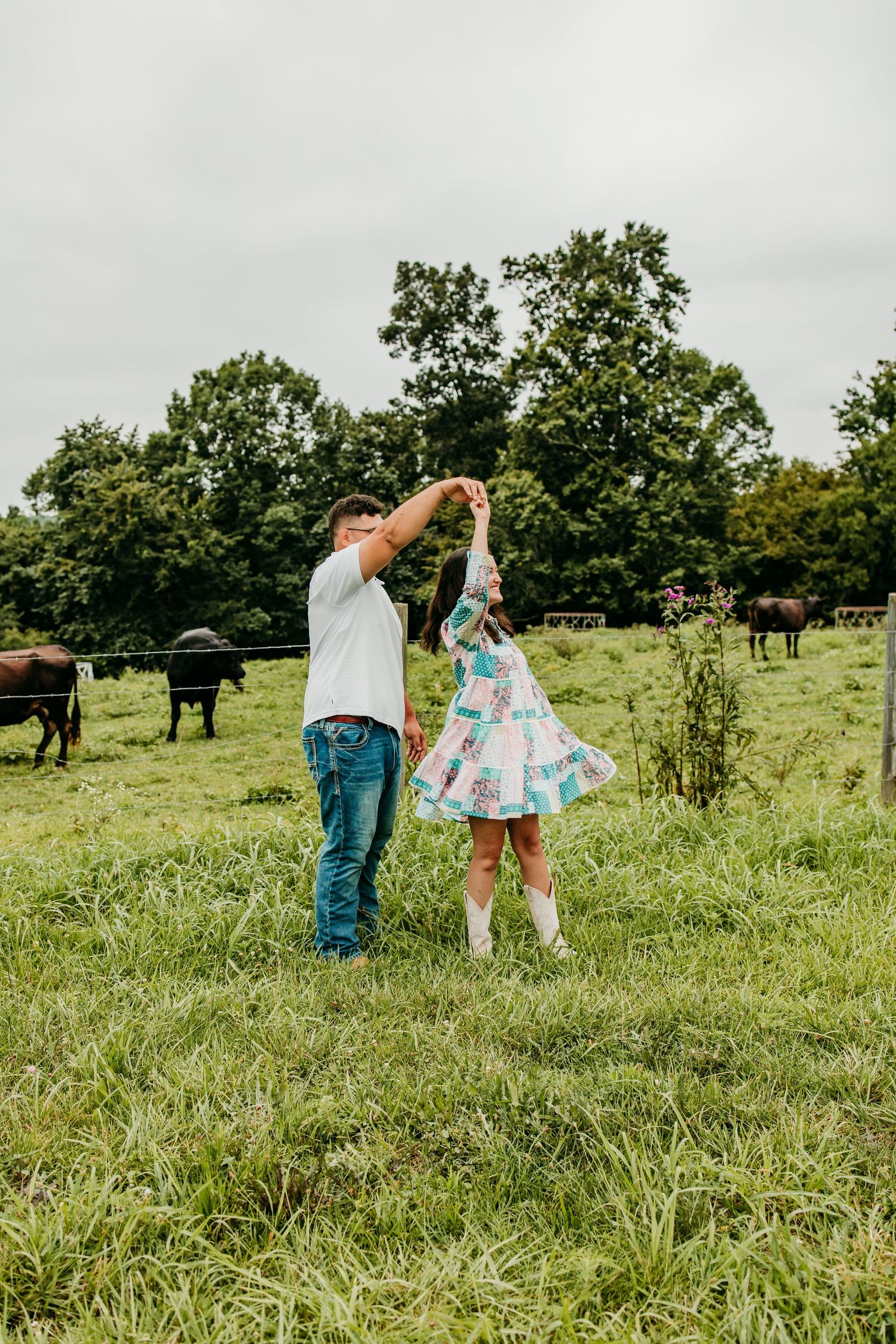 Couple dancing in a green pasture with cattle grazing in the background, woman wearing a floral dress and white boots.