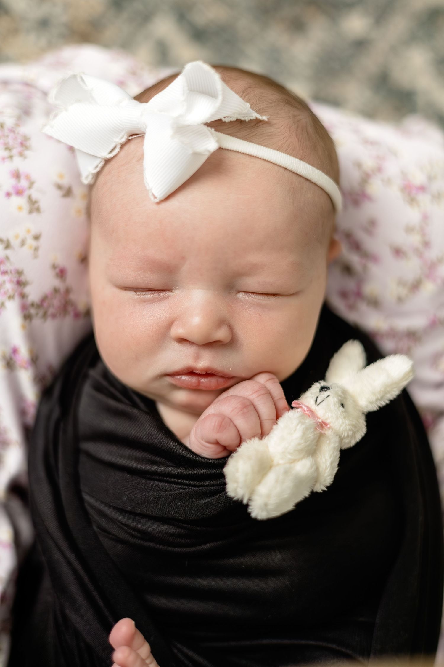 Sleeping newborn baby wrapped in black swaddle, wearing white bow headband, holding small white bunny stuffed animal.