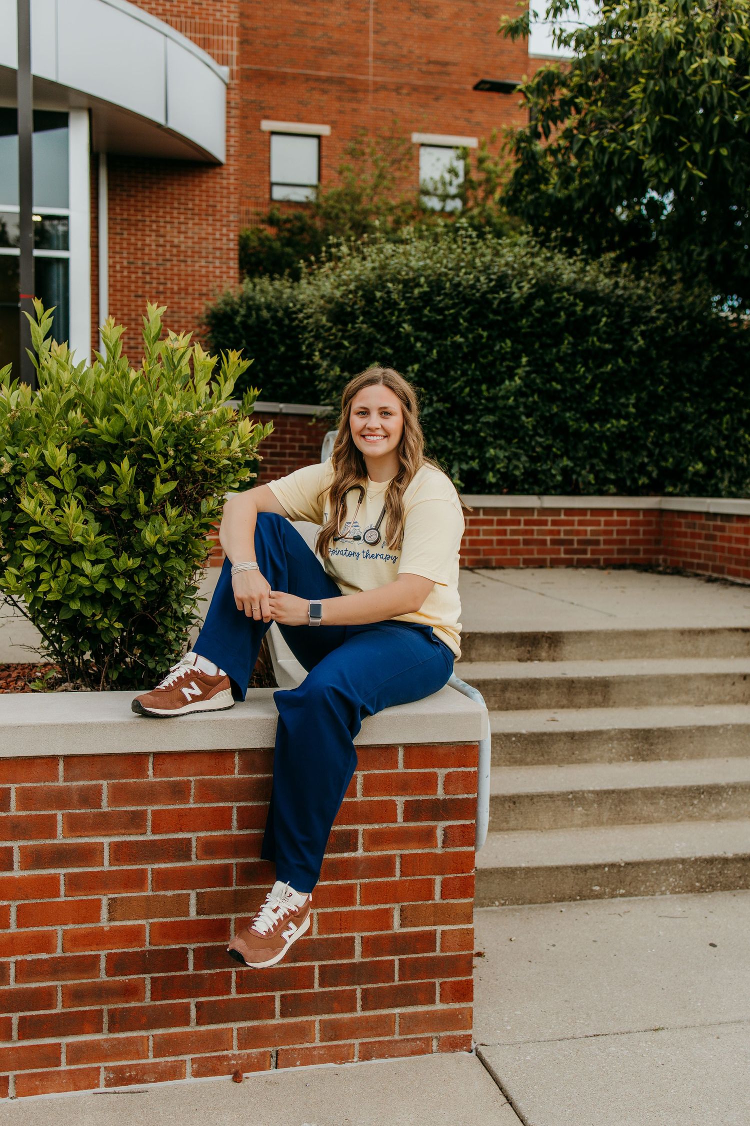Young woman smiling in blue pants and yellow tee, sitting on brick wall outside university building.