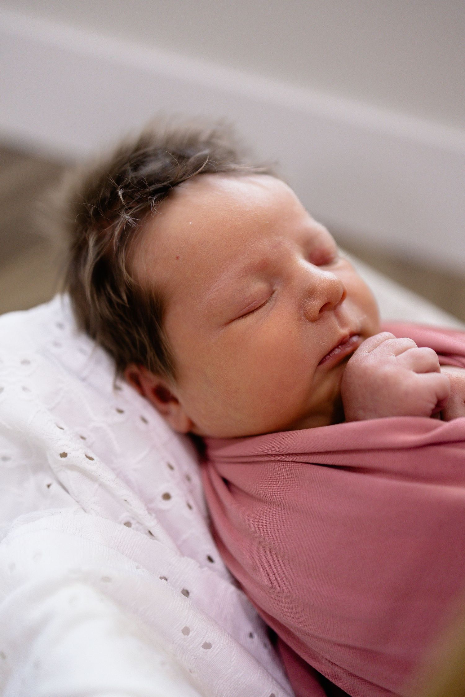 Sleeping newborn baby swaddled in a pink wrap, lying peacefully with dark hair and tiny hands visible.