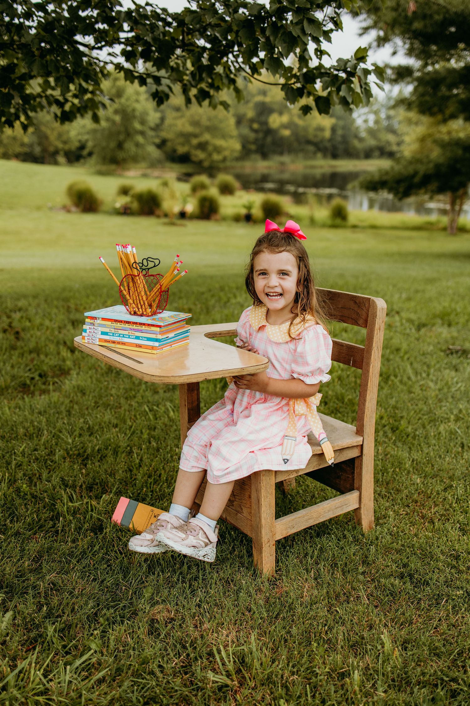 A smiling little girl in a pink dress sits at a wooden school desk outdoors, with books and pencils, wearing a pink bow.