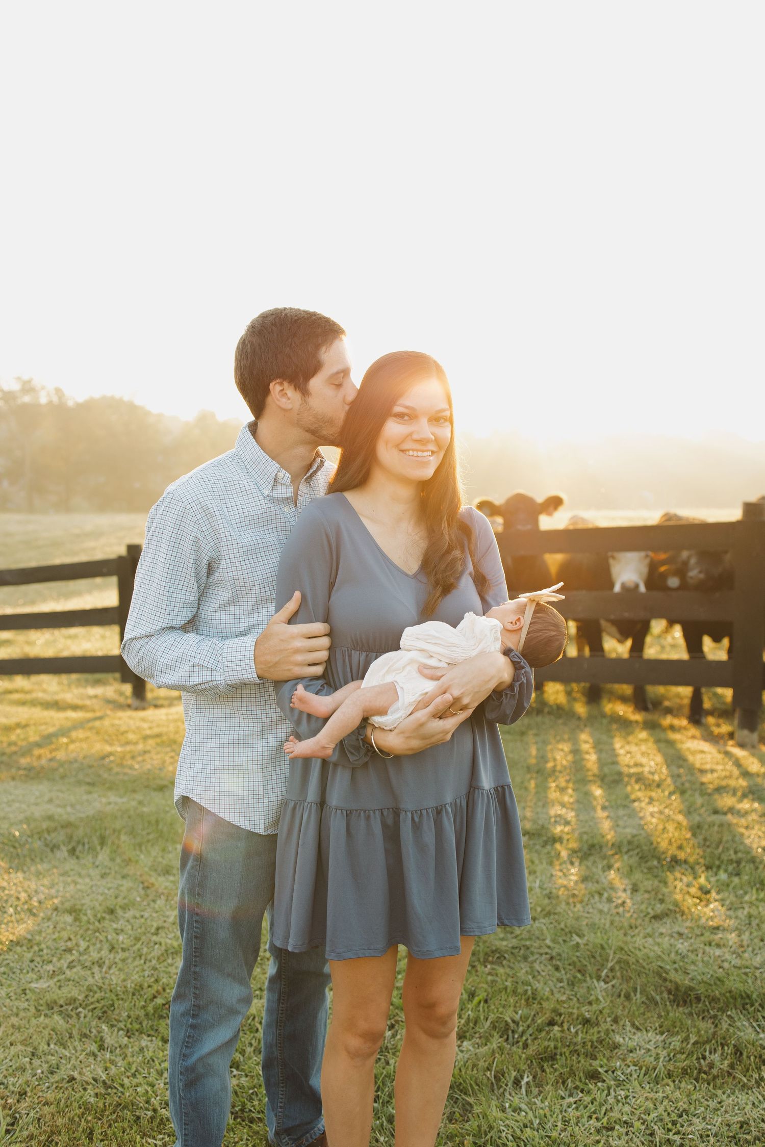 Young couple holding newborn baby outdoors at golden hour near a farm fence with cattle in background.