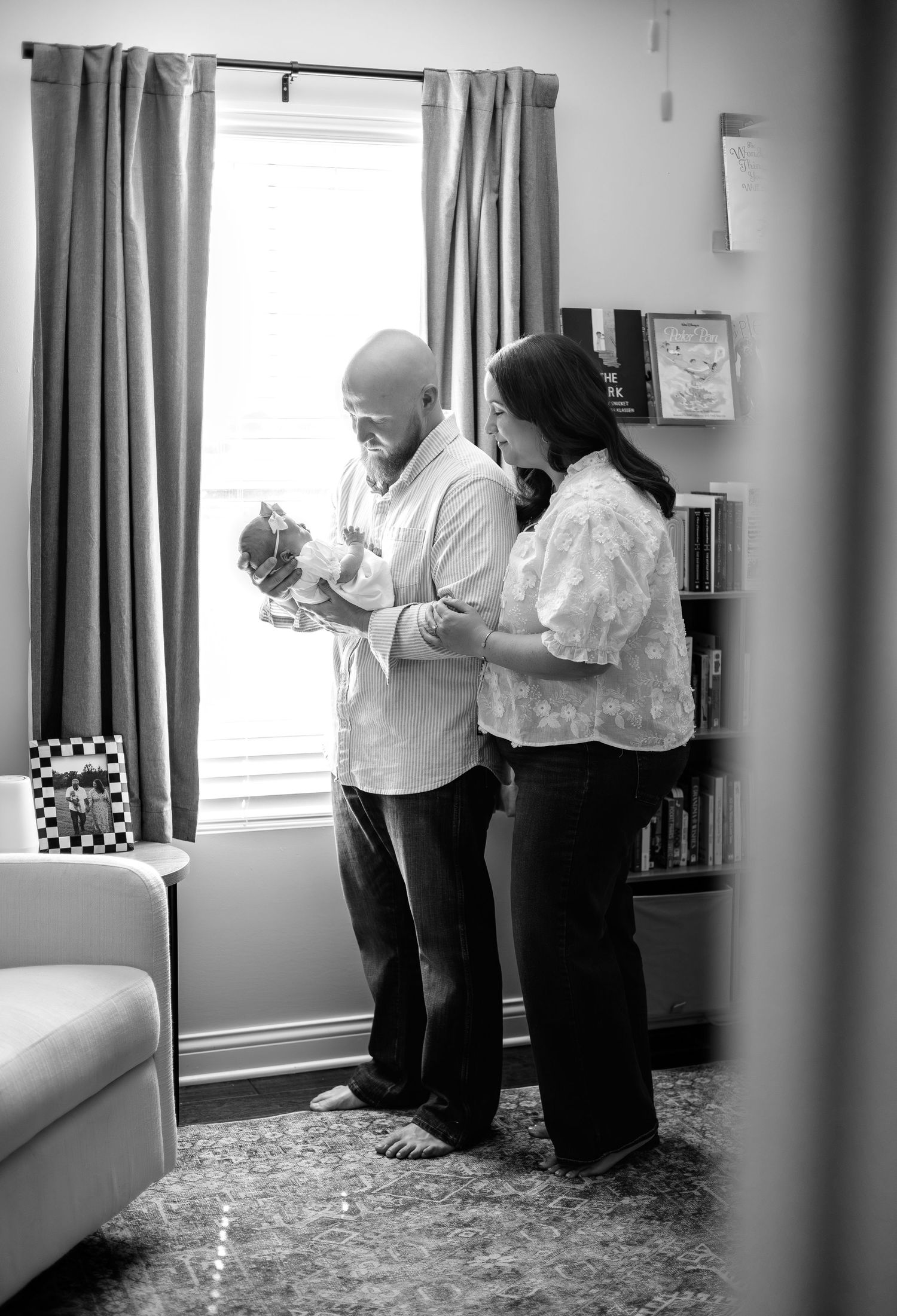 Black and white photo of a couple holding their newborn baby by a window in a cozy home setting.